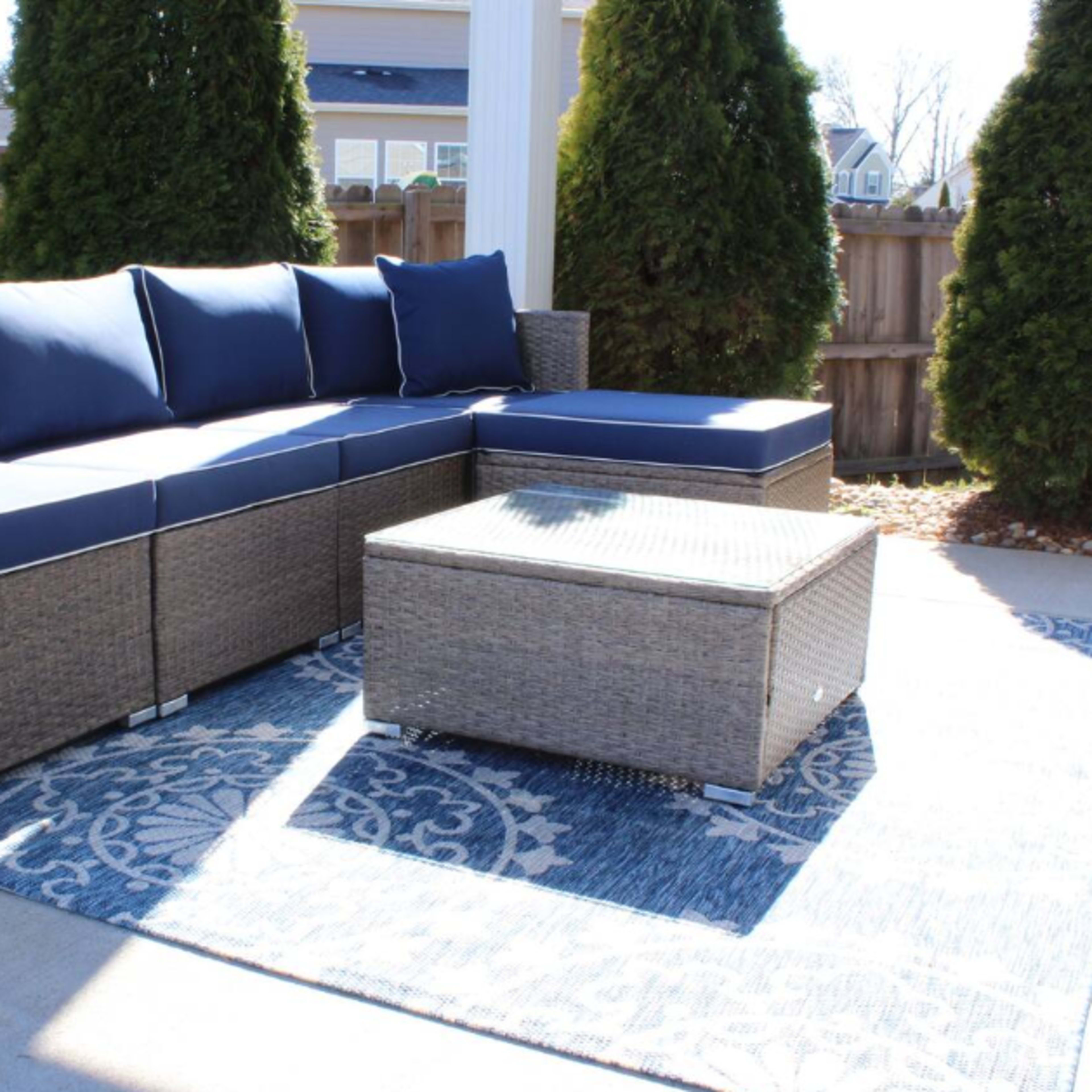 A sectional outdoor sofa with blue cushions and a matching coffee table is arranged on a patterned rug in a patio area surrounded by evergreen shrubs.