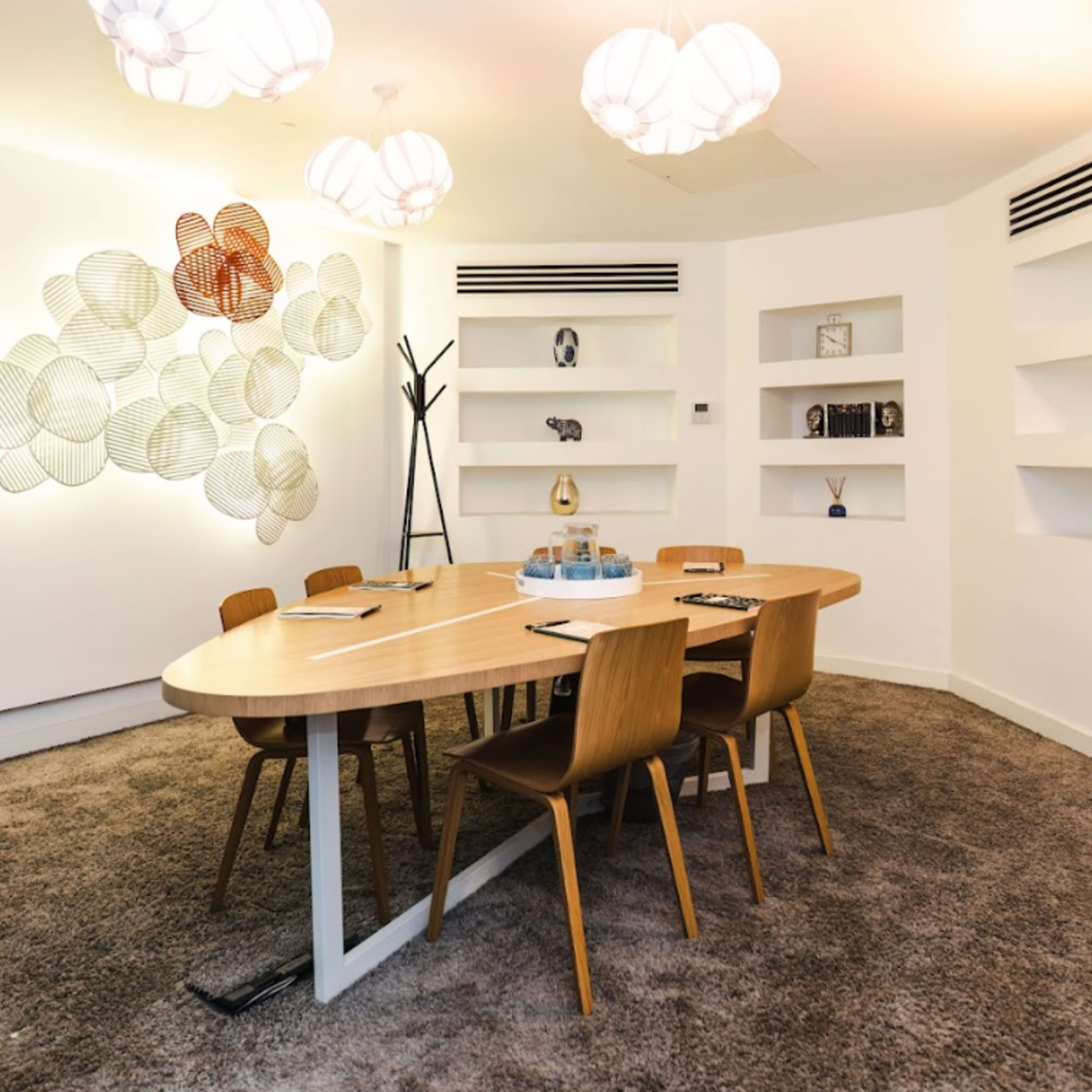 A modern conference room features a round wooden table surrounded by four wooden chairs, with decorative wall art and built-in shelves displaying various objects.