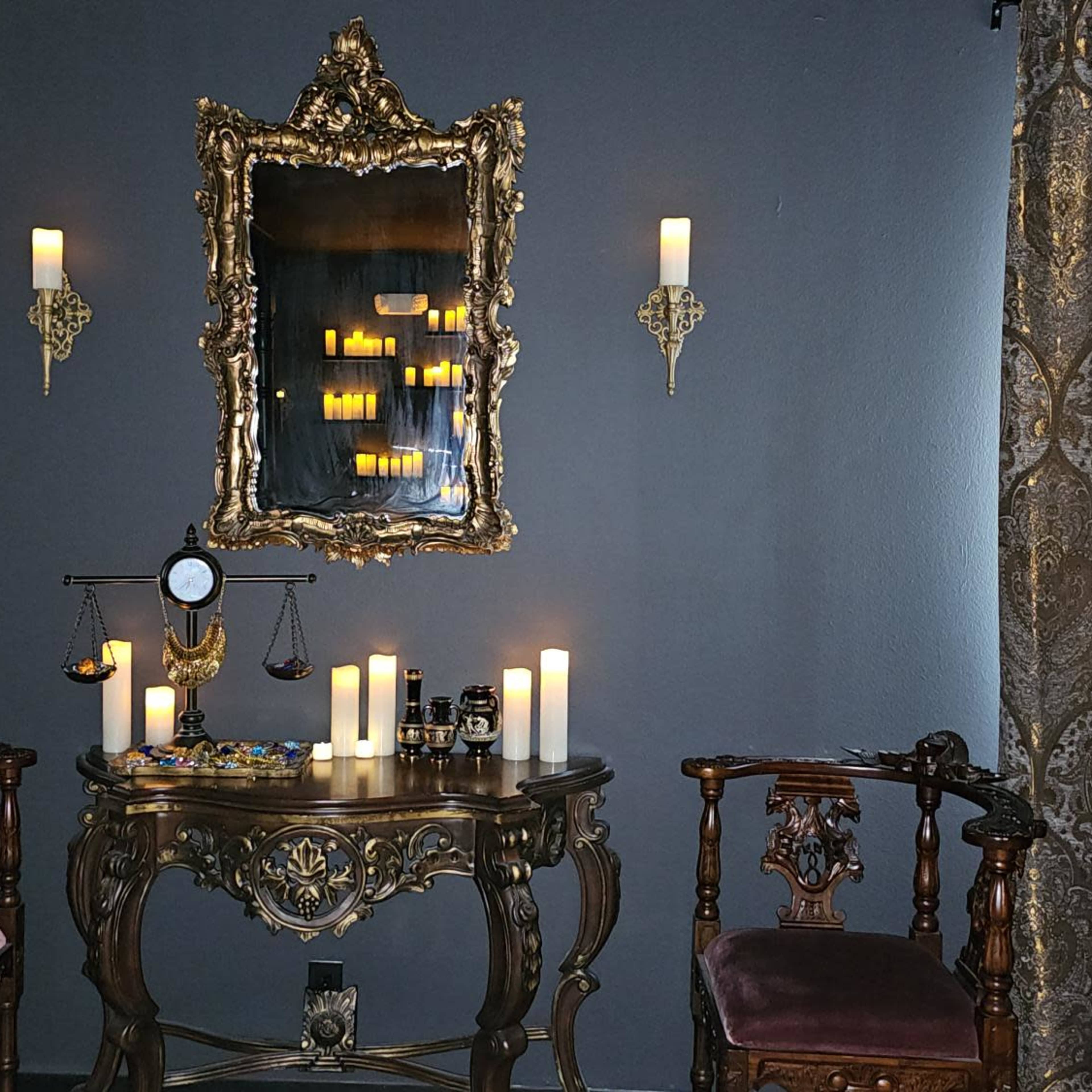 A dimly lit room featuring an ornate mirror surrounded by wall-mounted candles, a stylish wooden table with decorative items, and two vintage chairs.