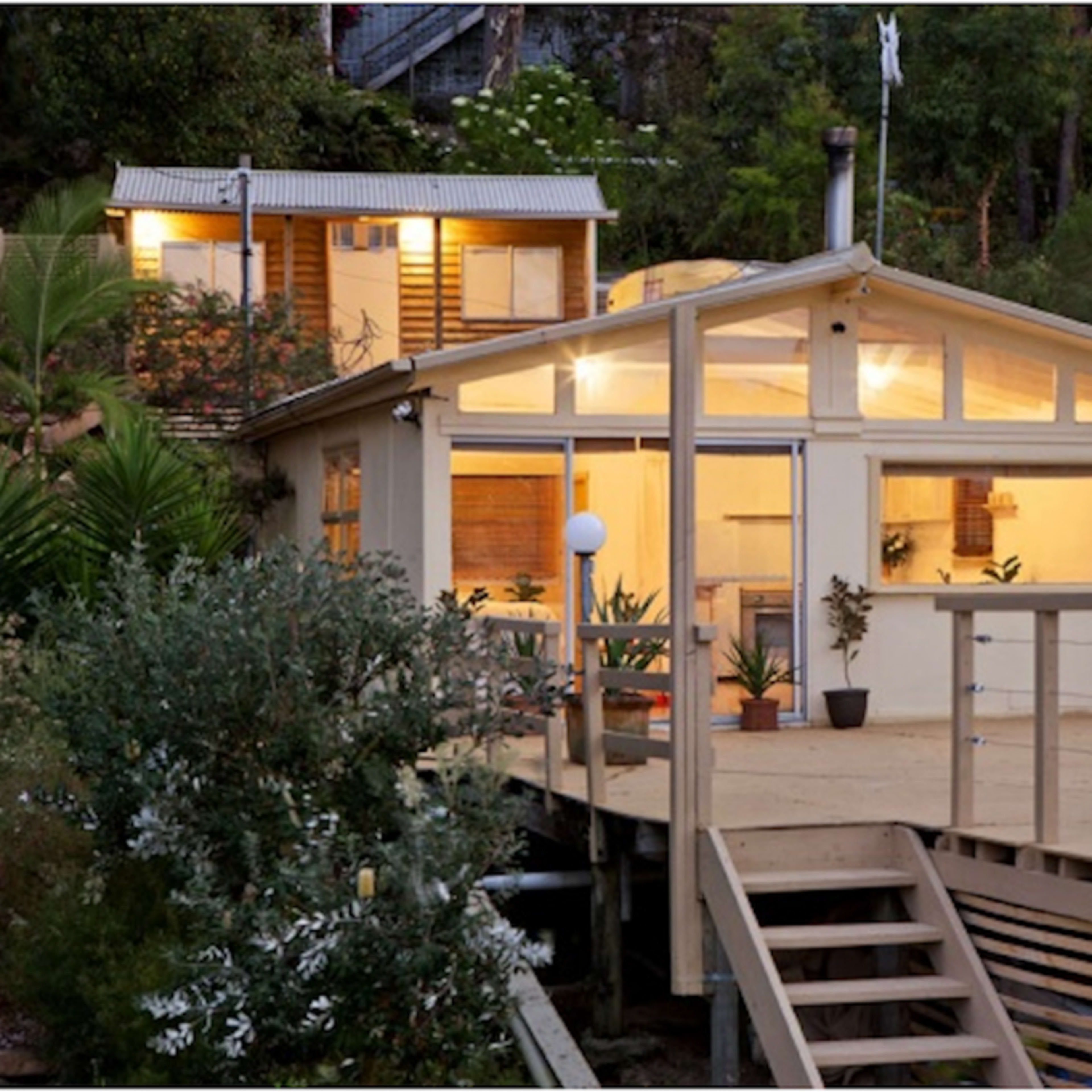 A wooden house with a large deck is situated on a hillside, showcasing large windows and surrounded by greenery.