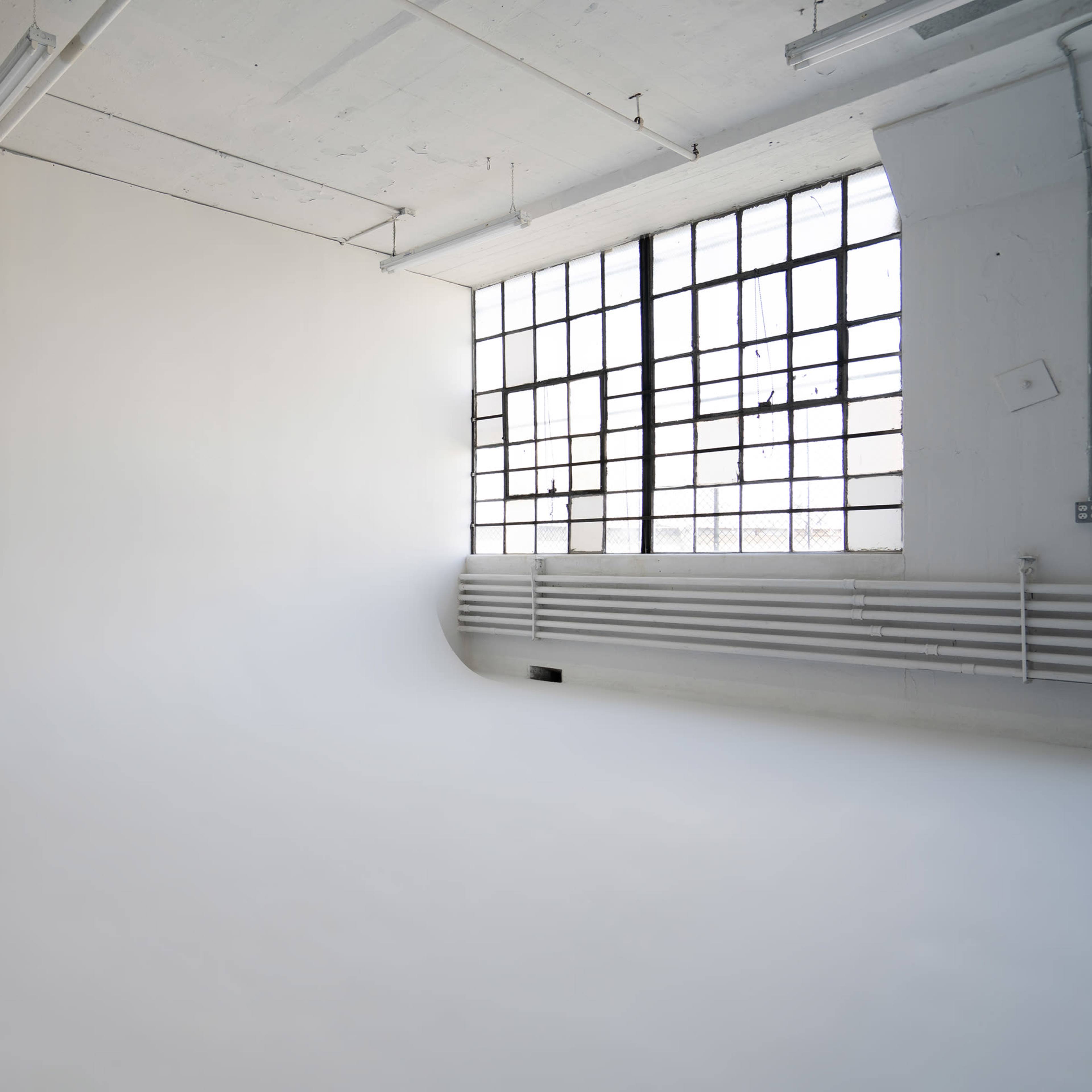 A spacious, empty studio features a seamless white backdrop and large industrial windows letting in natural light.