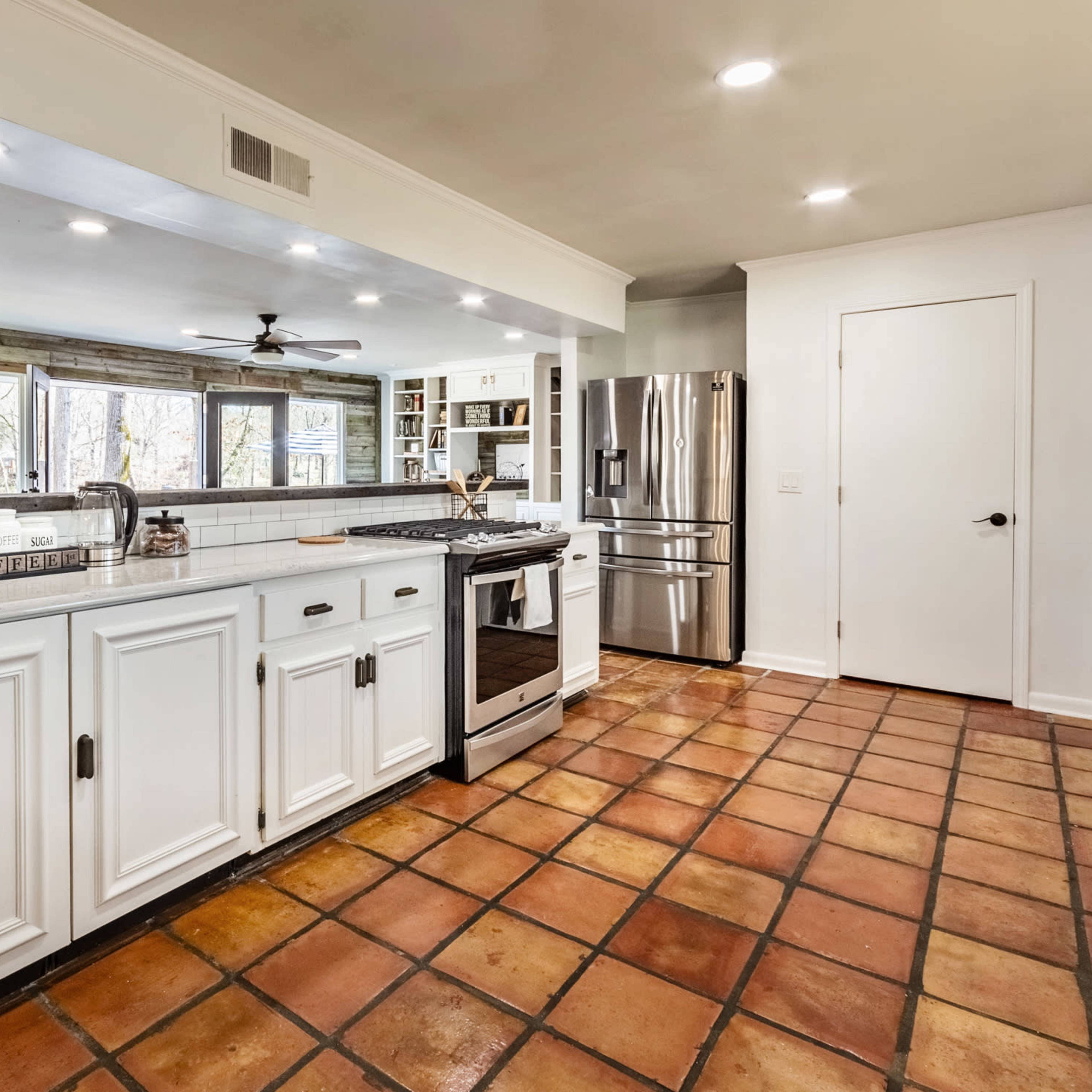 A modern kitchen features white cabinetry, a tiled floor, and stainless steel appliances, with large windows allowing natural light.