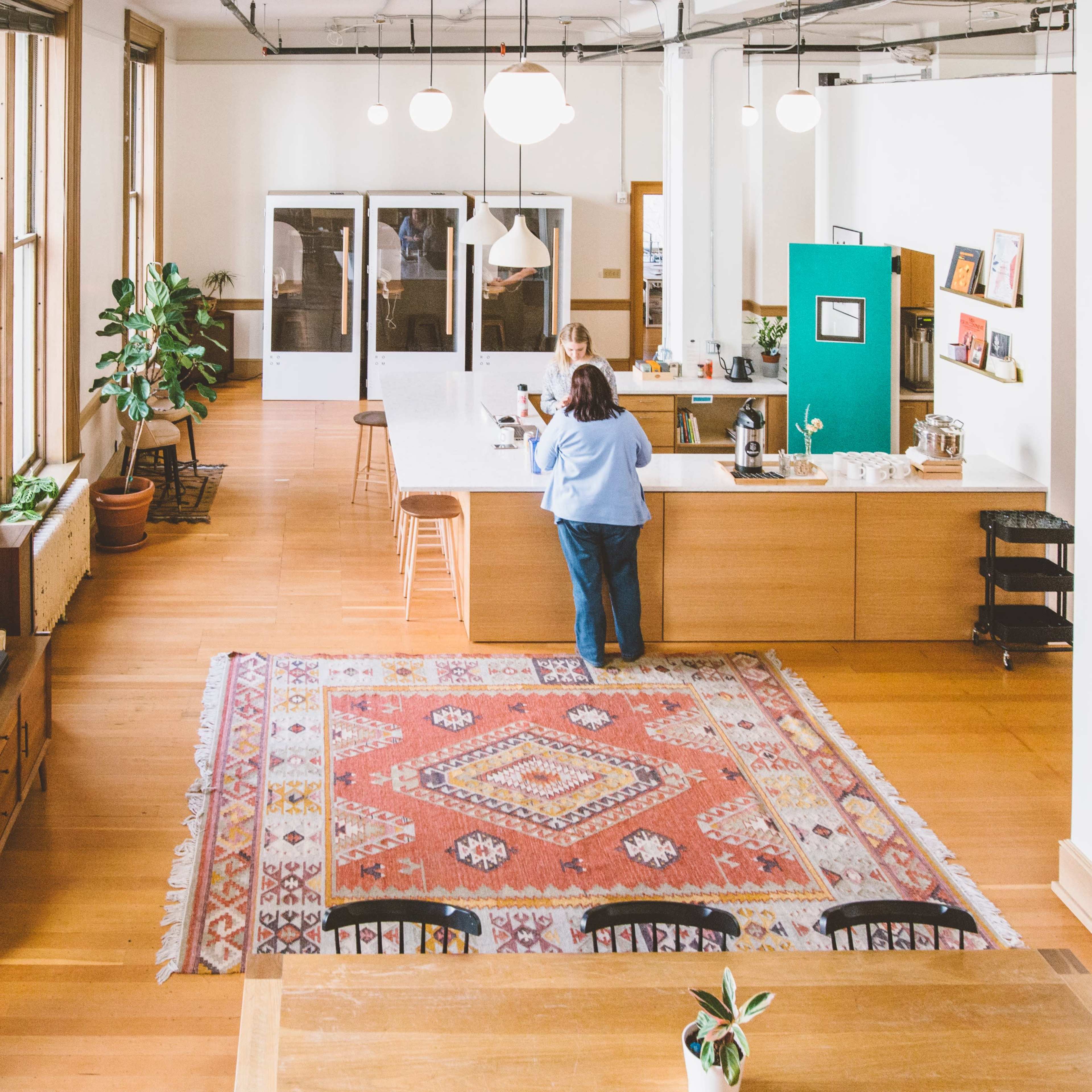 A spacious office interior features a large area rug, plants, a coffee station, and a person standing at a wooden counter.