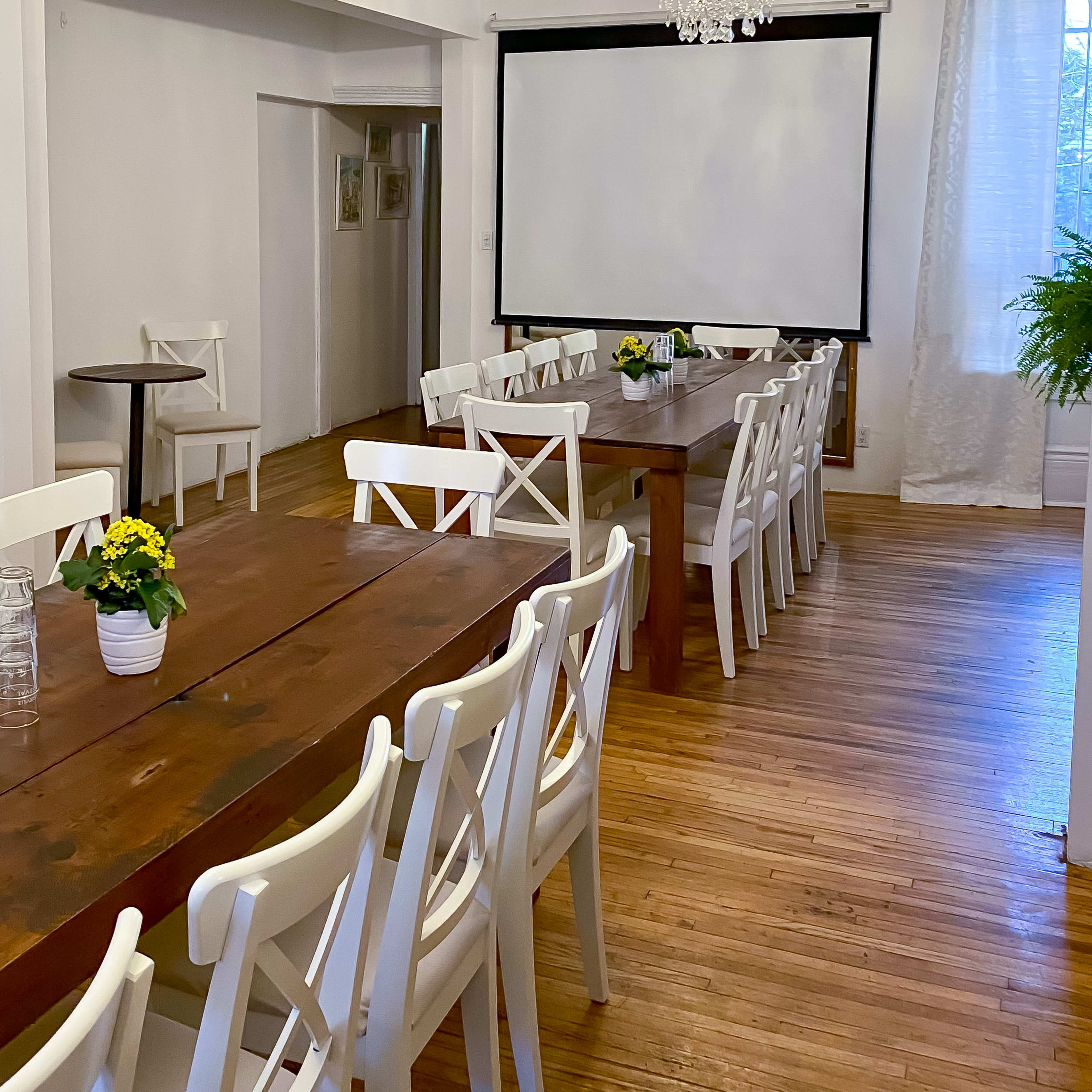 A meeting room features two long wooden tables surrounded by white chairs, with a large screen at one end and a chandelier overhead.