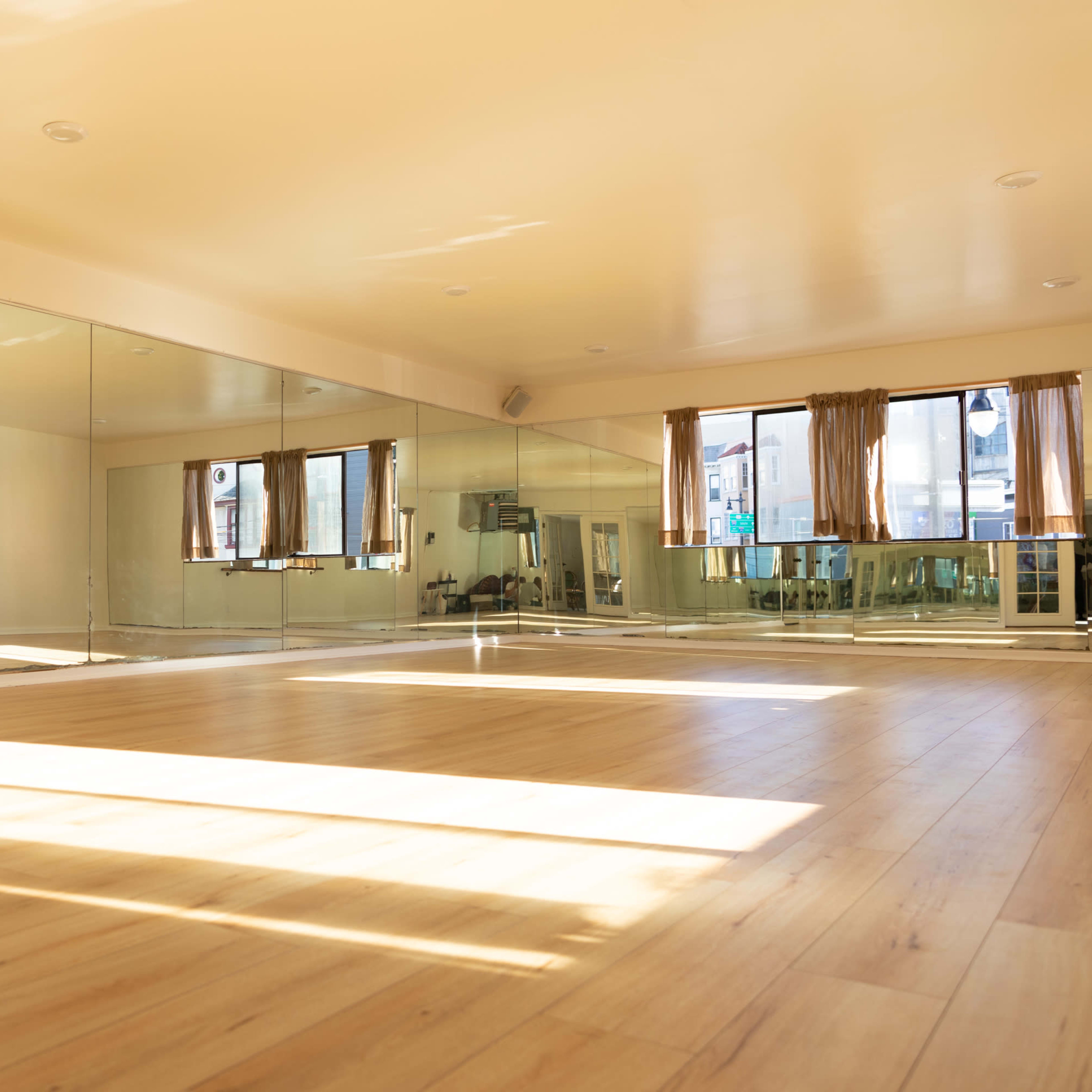 A spacious dance studio features large mirrors, wooden flooring, and natural light streaming through windows with sheer curtains.