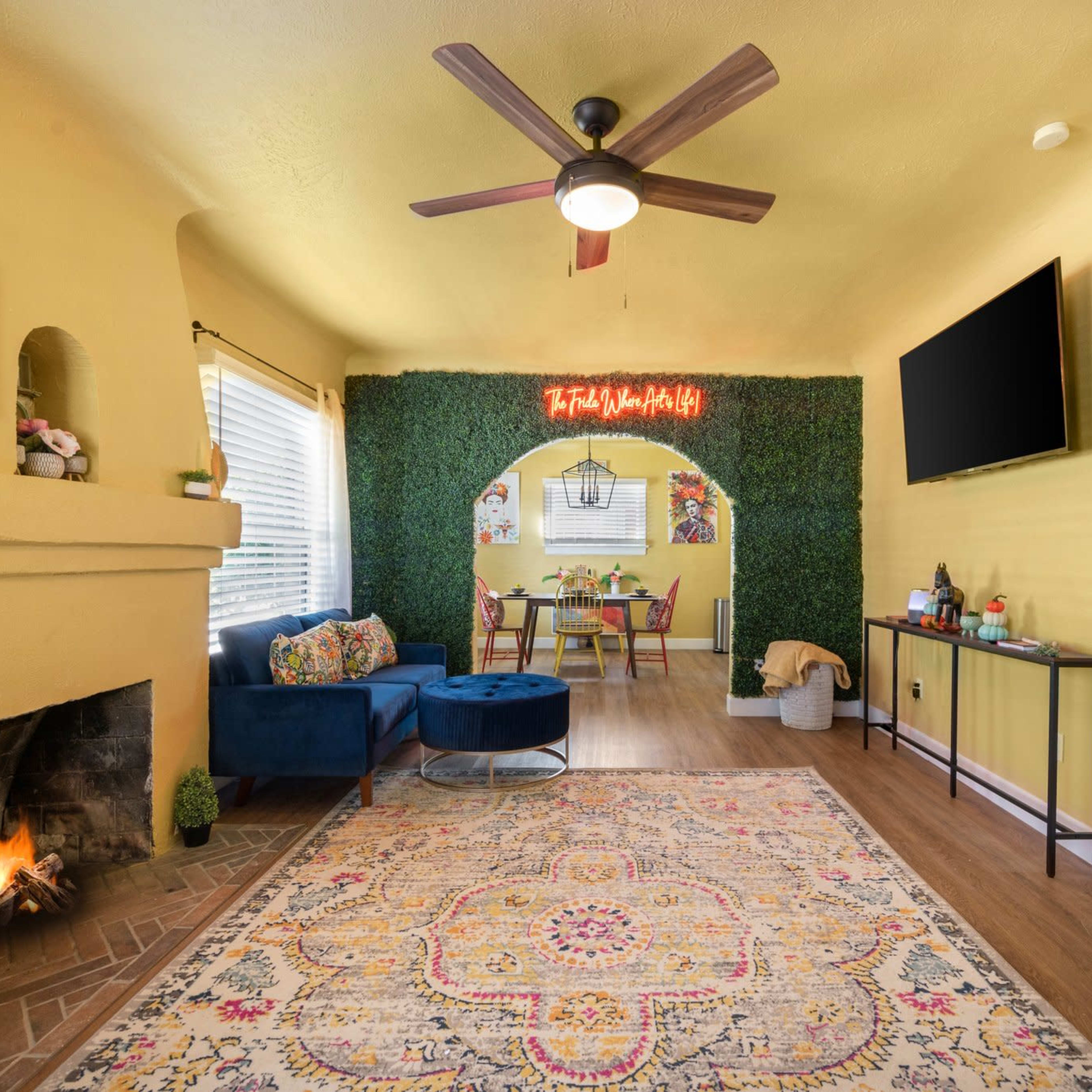 A brightly colored living room featuring a fireplace, a blue seating area, and a dining space with a green wall decoration and a TV mounted on the wall.
