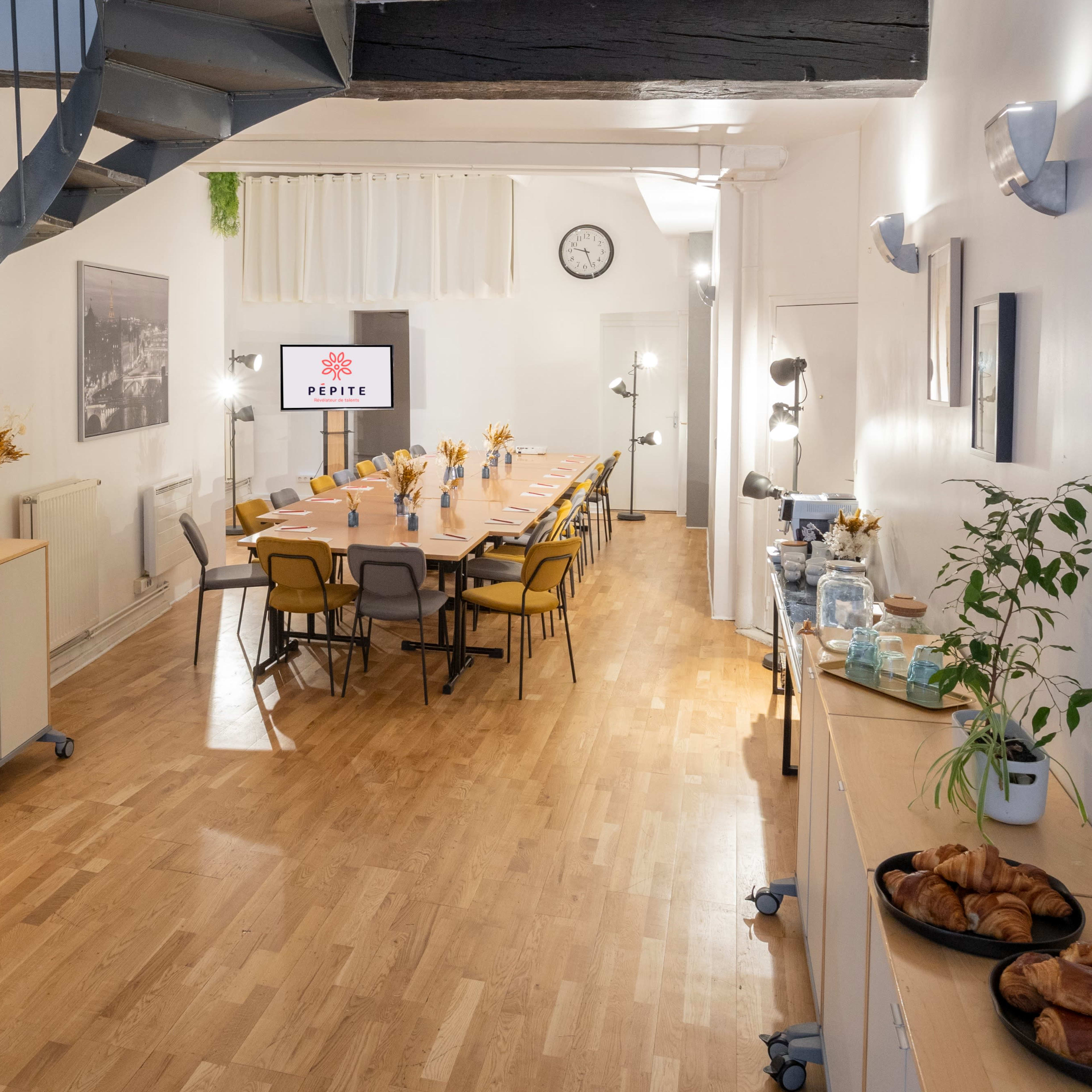 A well-lit, modern meeting room with a long table set for an event, featuring pastries and beverages along the walls, and a staircase in the background.