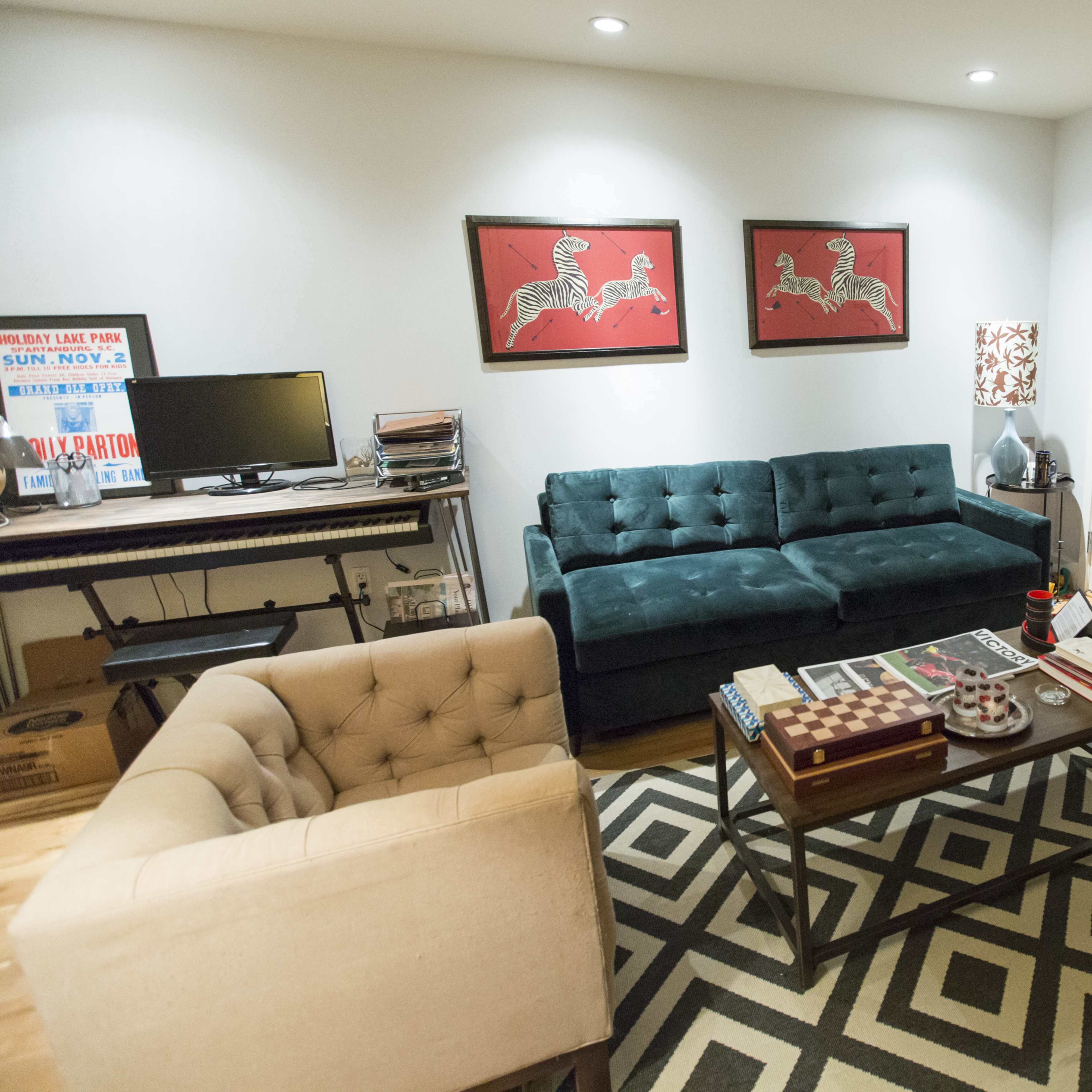 A small living room with a green tufted couch, a beige armchair, a coffee table with books and a decorative item, and two red artwork pieces on the wall.