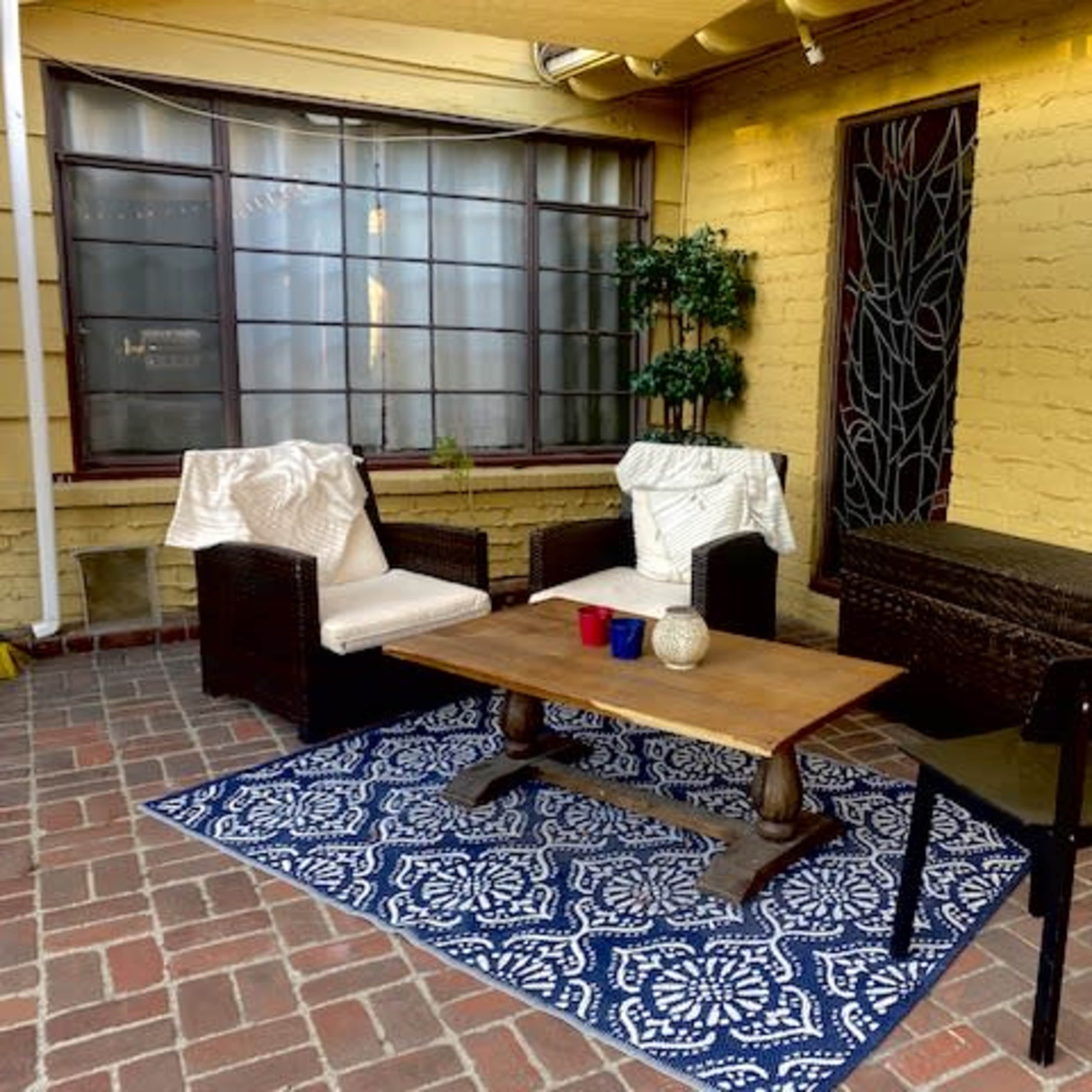 A small outdoor seating area with two wicker chairs, a wooden coffee table, a patterned rug, and a potted plant against a yellow brick wall.