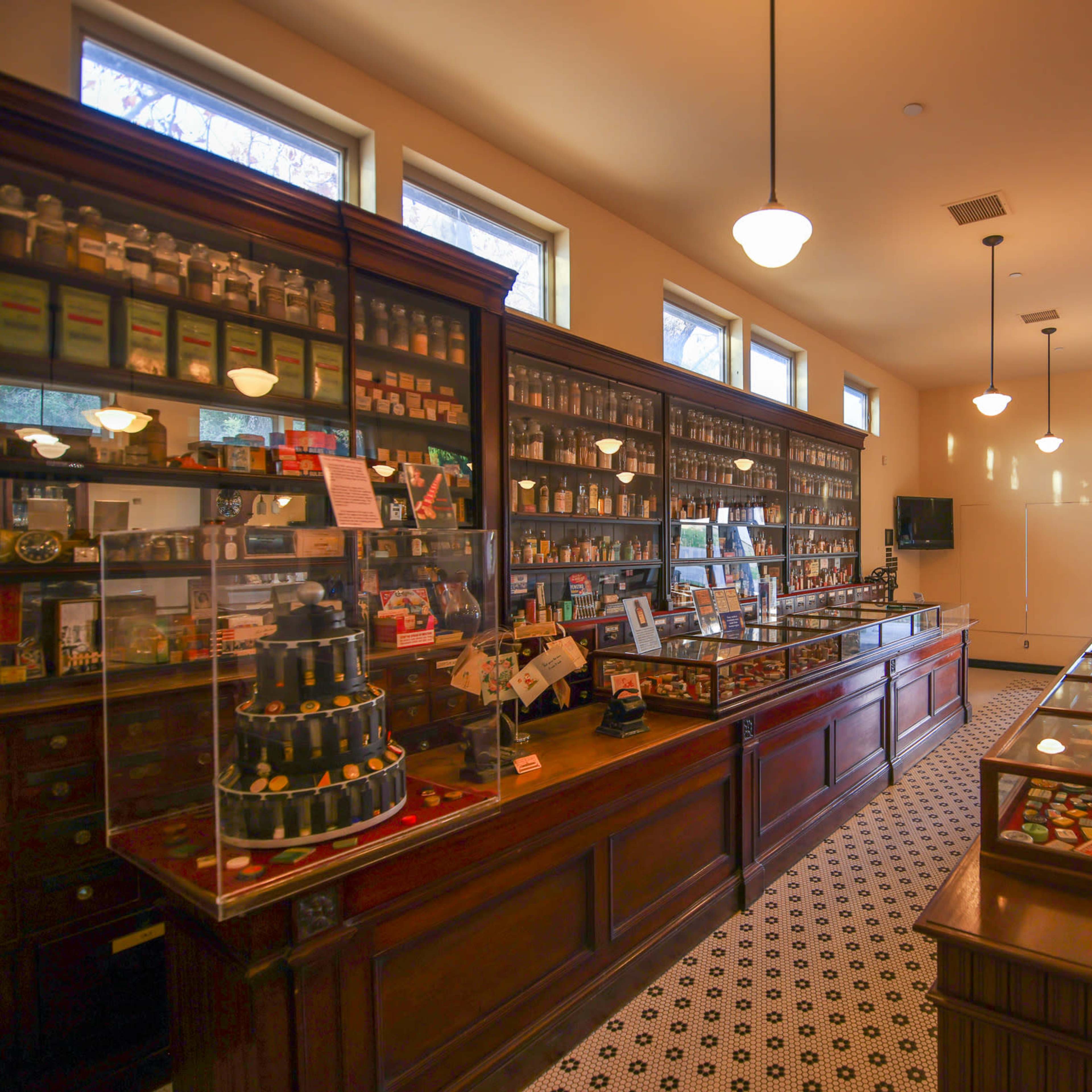 A vintage apothecary shop interior with wooden displays, glass cases filled with jars and various items, and large windows along one wall.