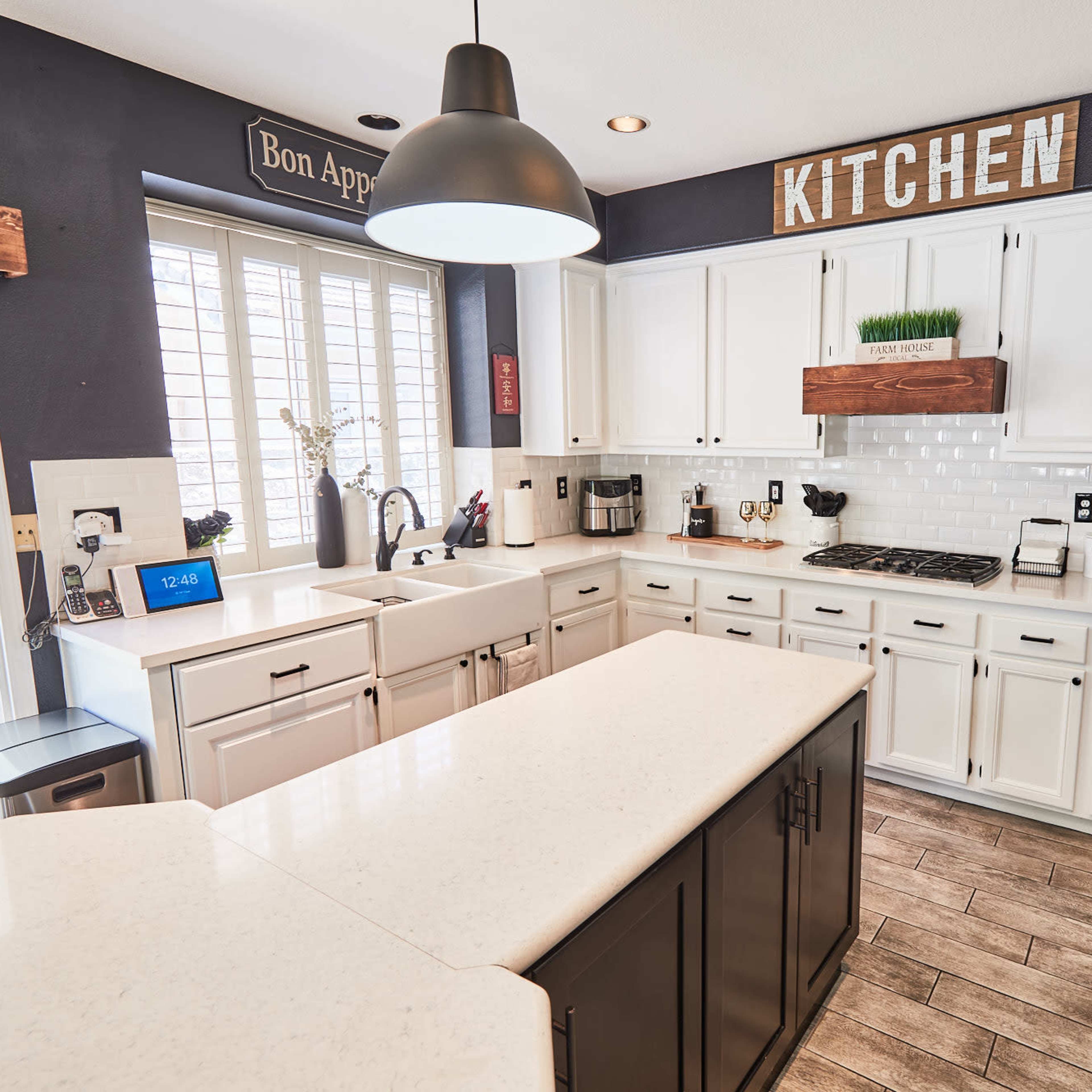 A modern kitchen features white cabinets, a central island, and large windows with natural light illuminating the space.