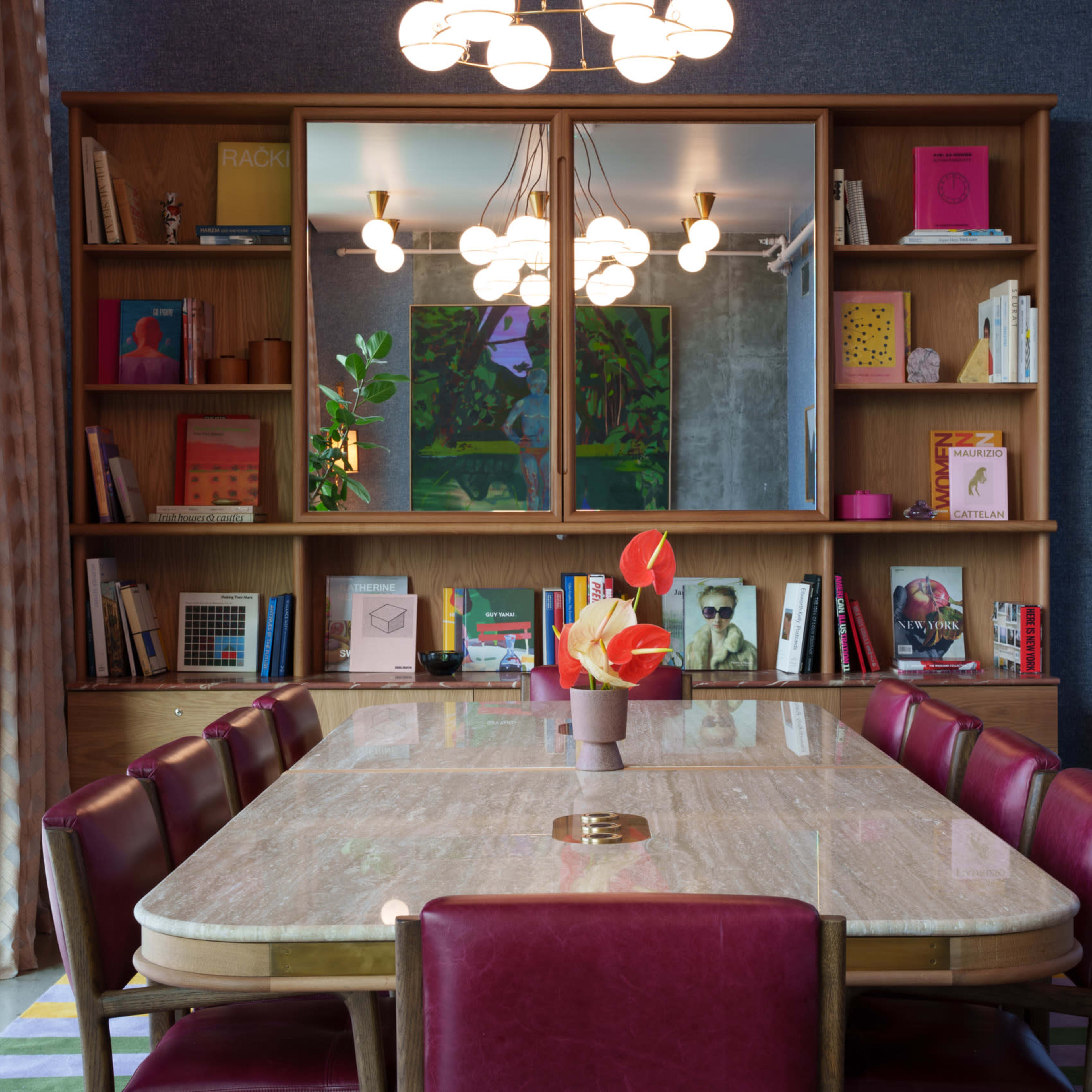 A modern dining room with a large table surrounded by burgundy chairs, a bookshelf filled with colorful books in the background, and a decorative arrangement of flowers on the table.