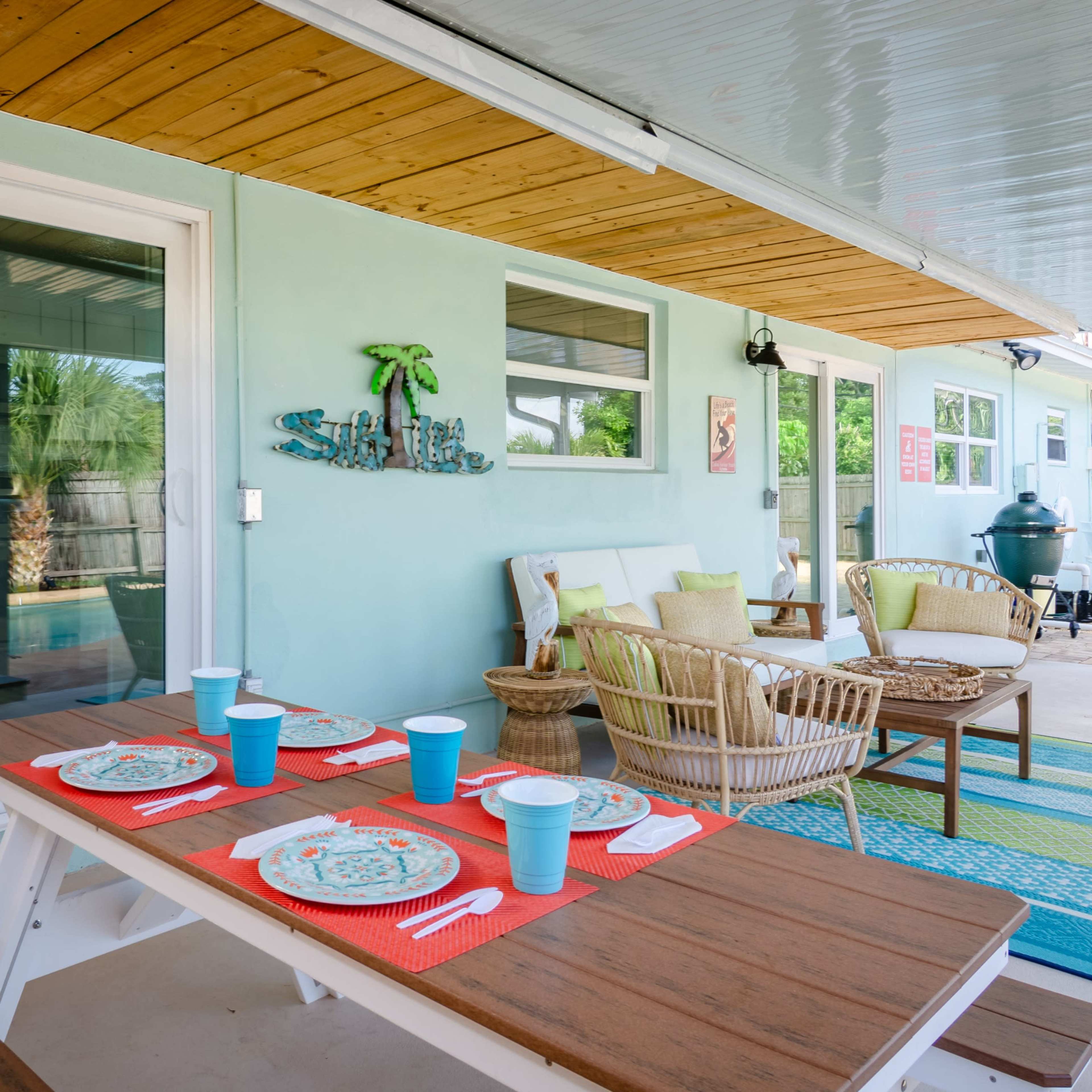 A patio area is set up with a table and chairs, featuring dishes and blue cups, next to a swimming pool and surrounded by greenery.