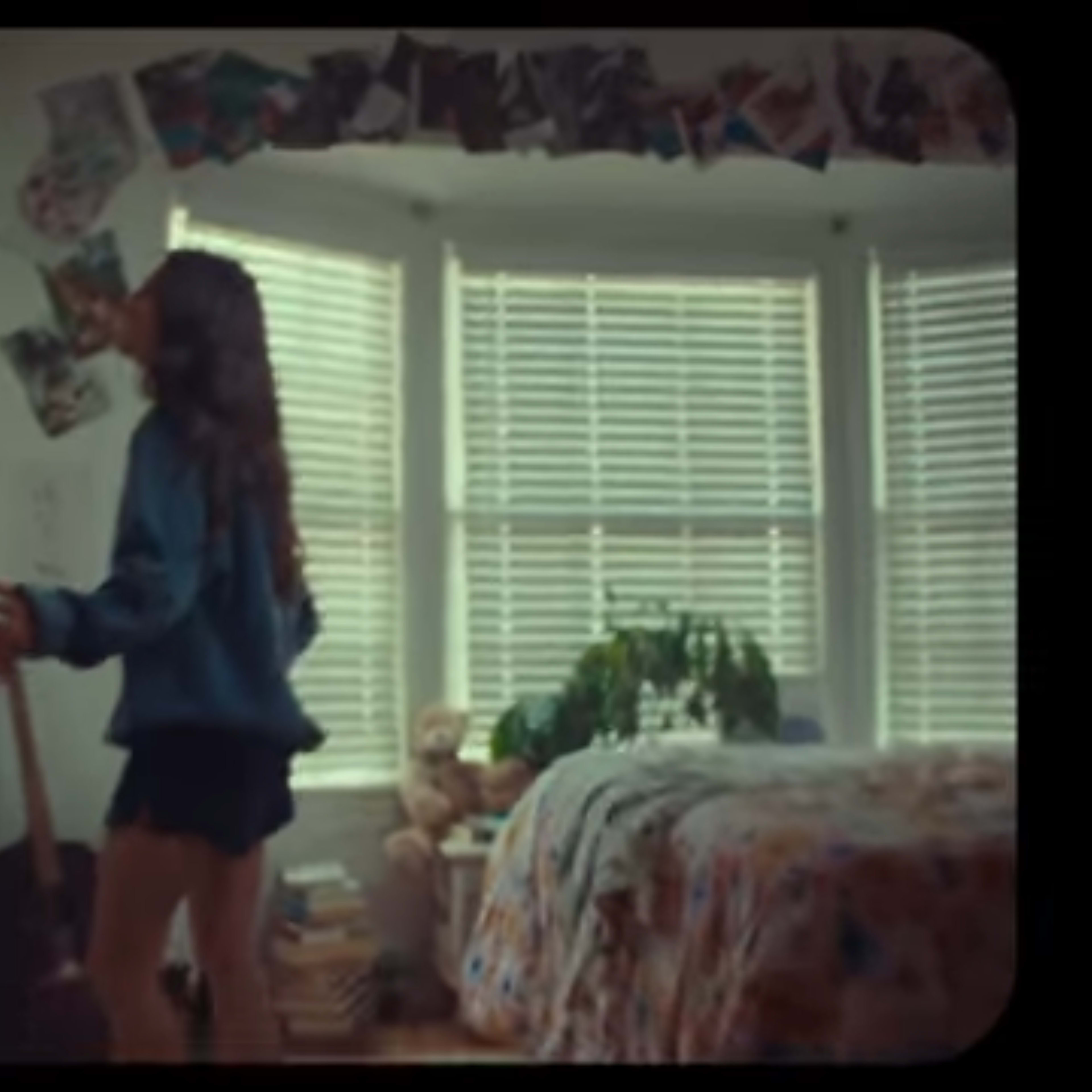 A person stands near a wall covered in photographs and sketches in a bright room with a bed and potted plants.