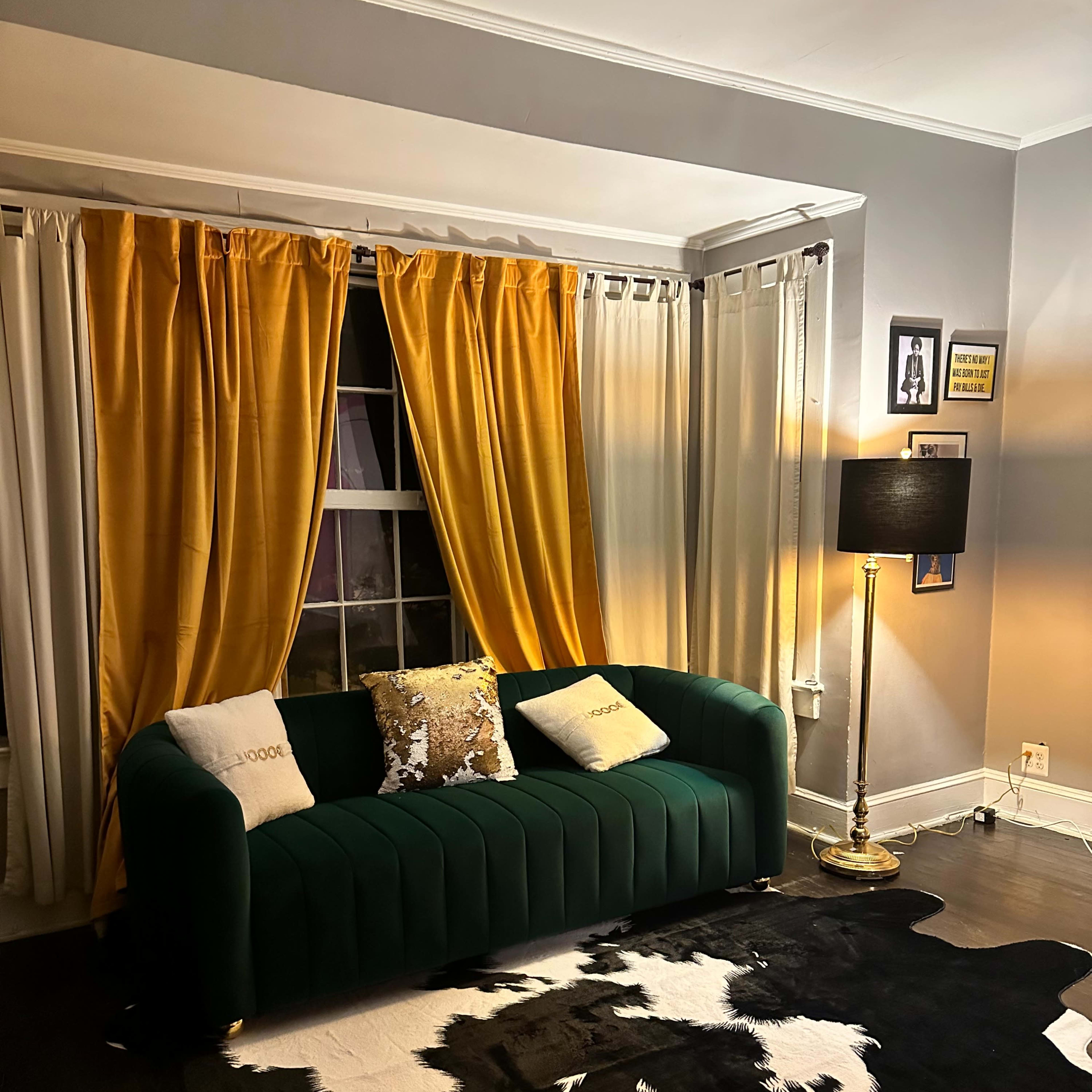 A green sofa is positioned in a living room with yellow and white curtains, a gold floor lamp, and a cowhide rug on a dark wood floor.