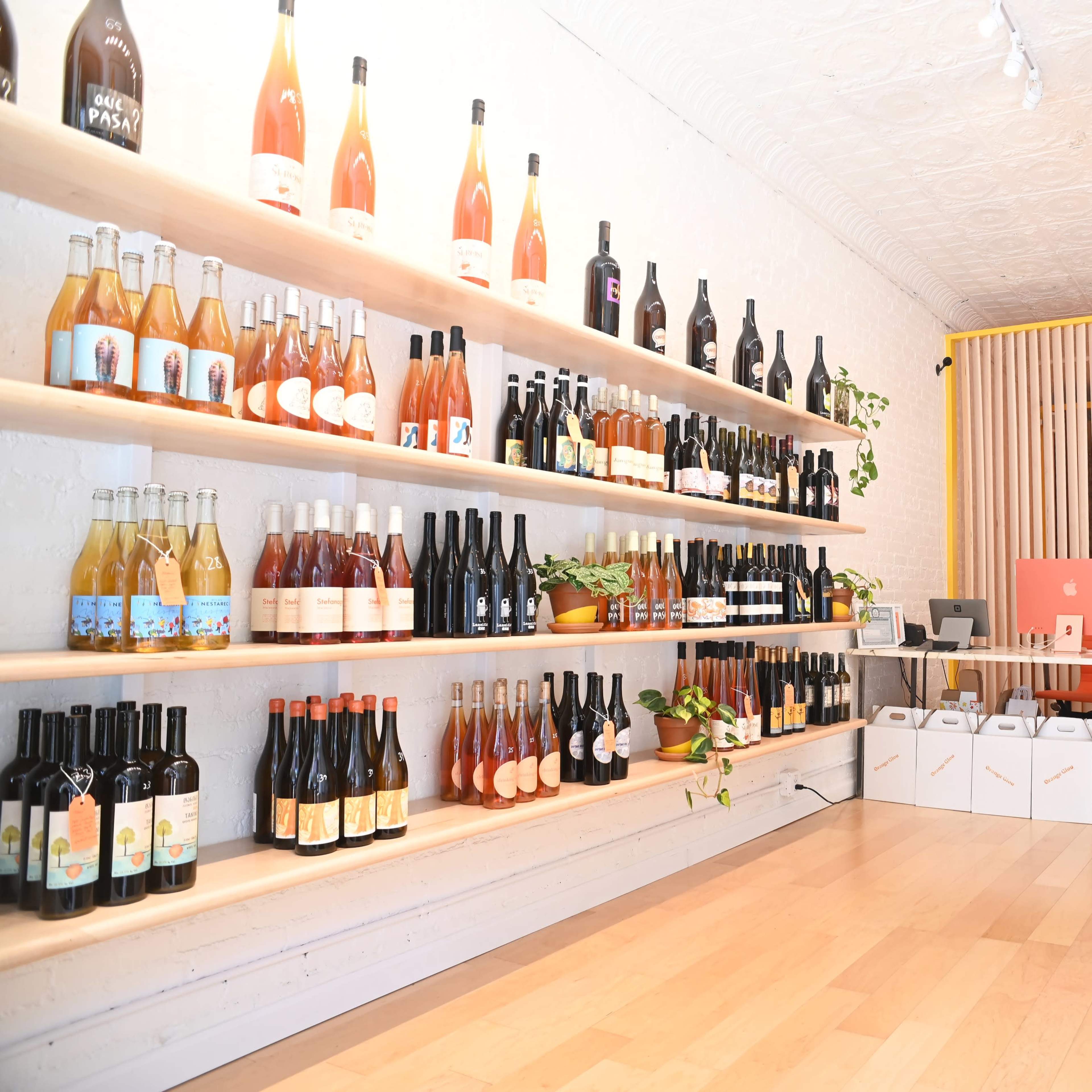A modern wine shop with a wooden shelf displaying various bottles of wine, and a bright seating area in the background featuring a colorful refrigerator and partition.