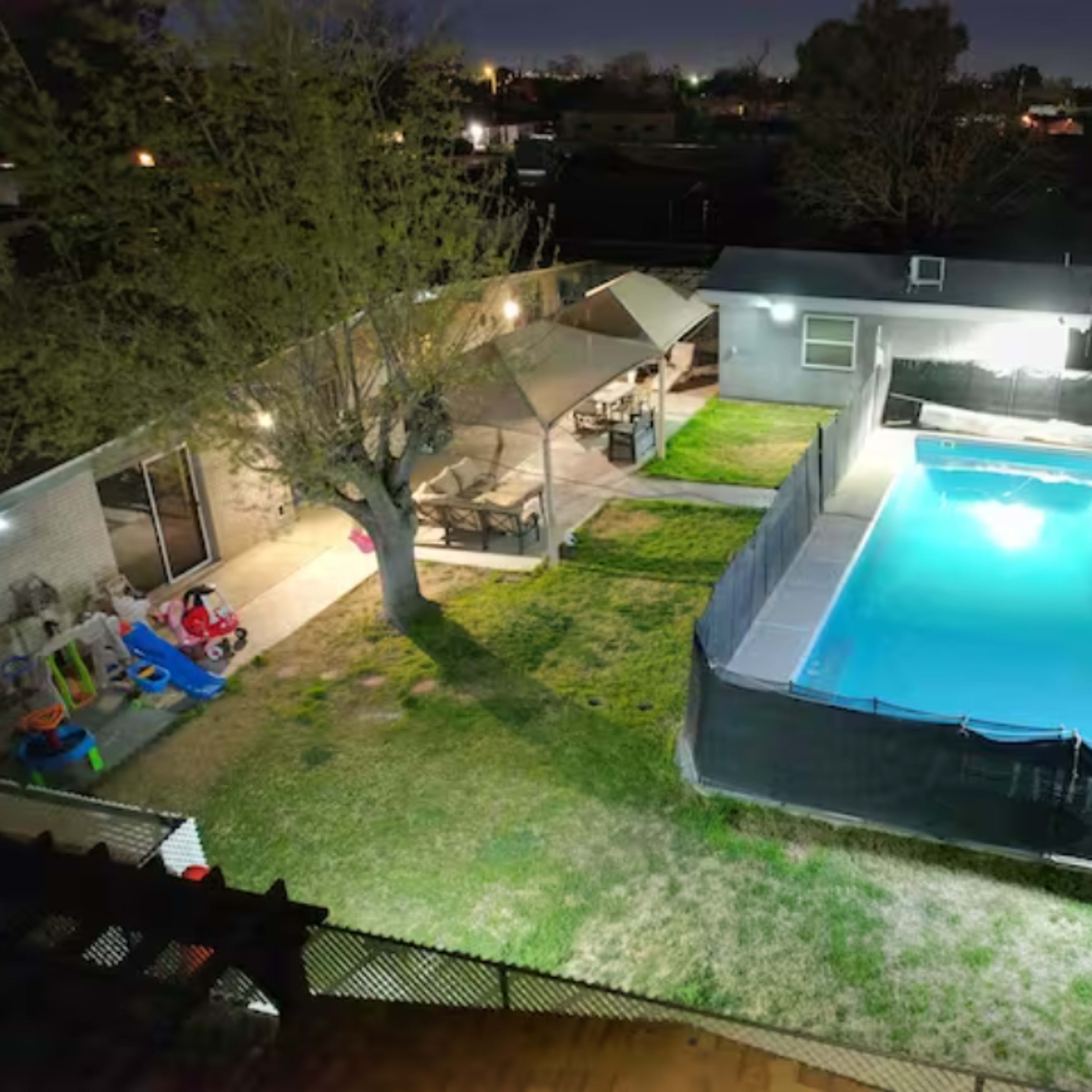 A backyard scene at night featuring a swimming pool surrounded by grass, with a seating area and play equipment nearby.
