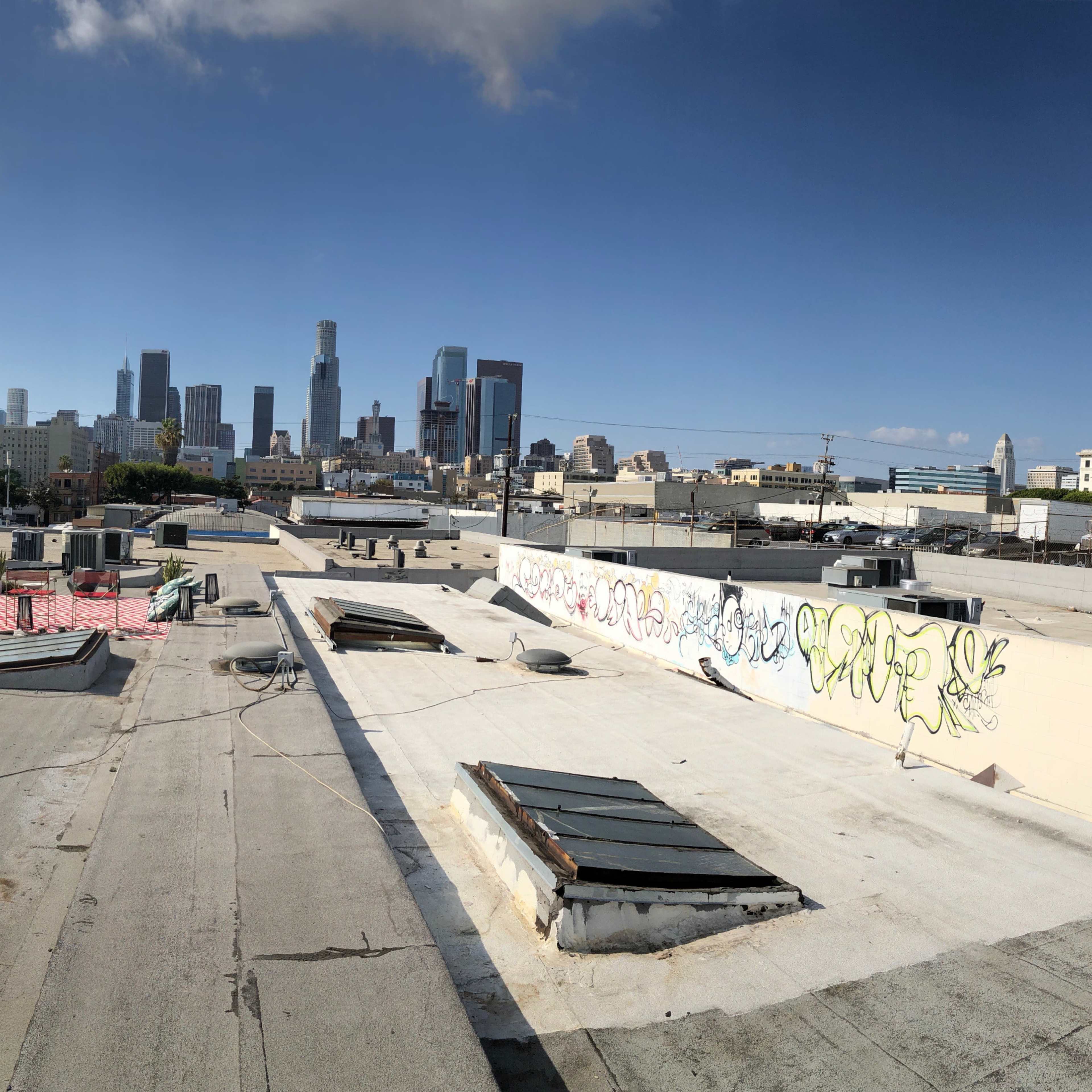 A panoramic view of rooftops in the foreground with the skyline of downtown Los Angeles in the background under a partly cloudy sky.