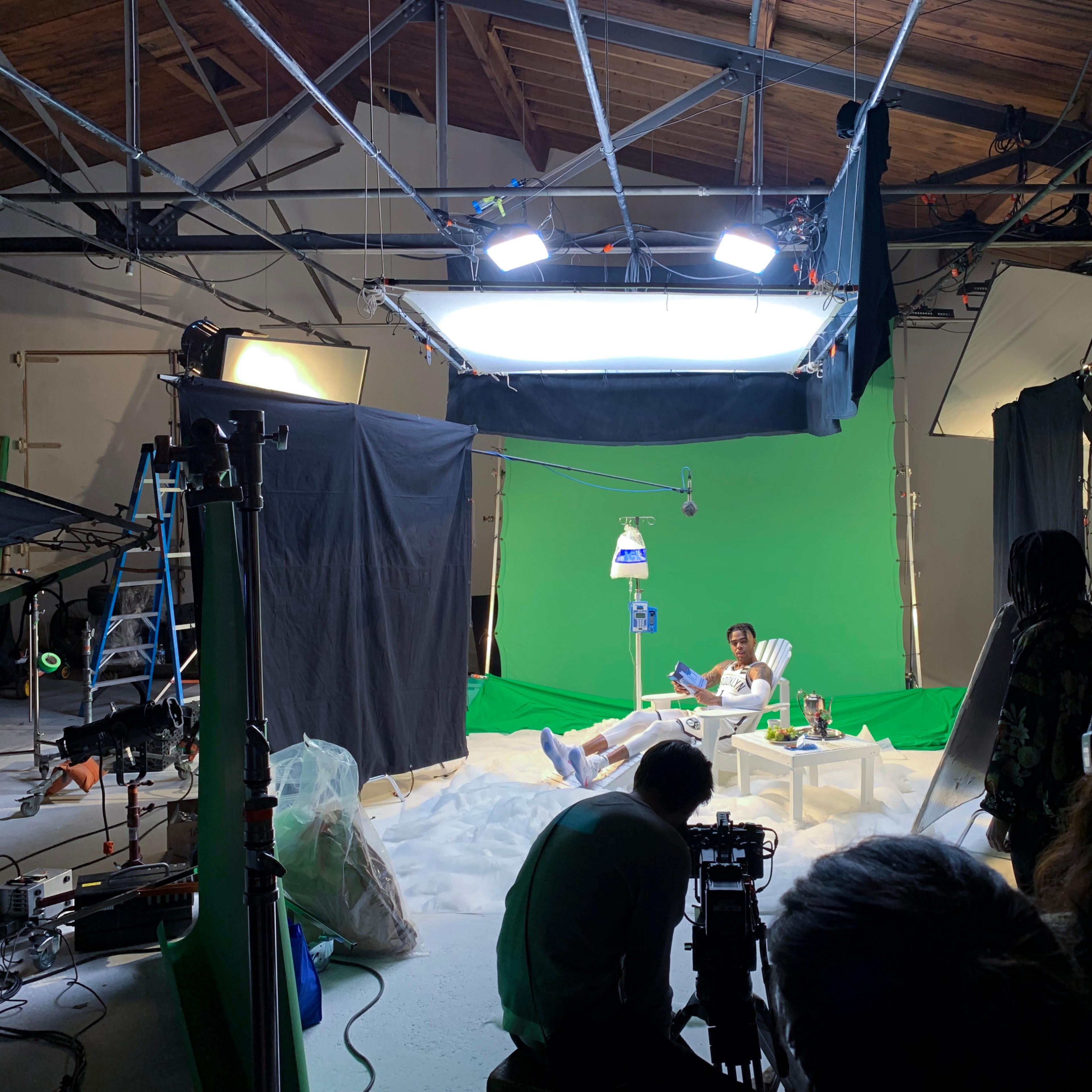 A film set features a green screen backdrop with a person sitting on a white couch surrounded by production equipment and crew members.