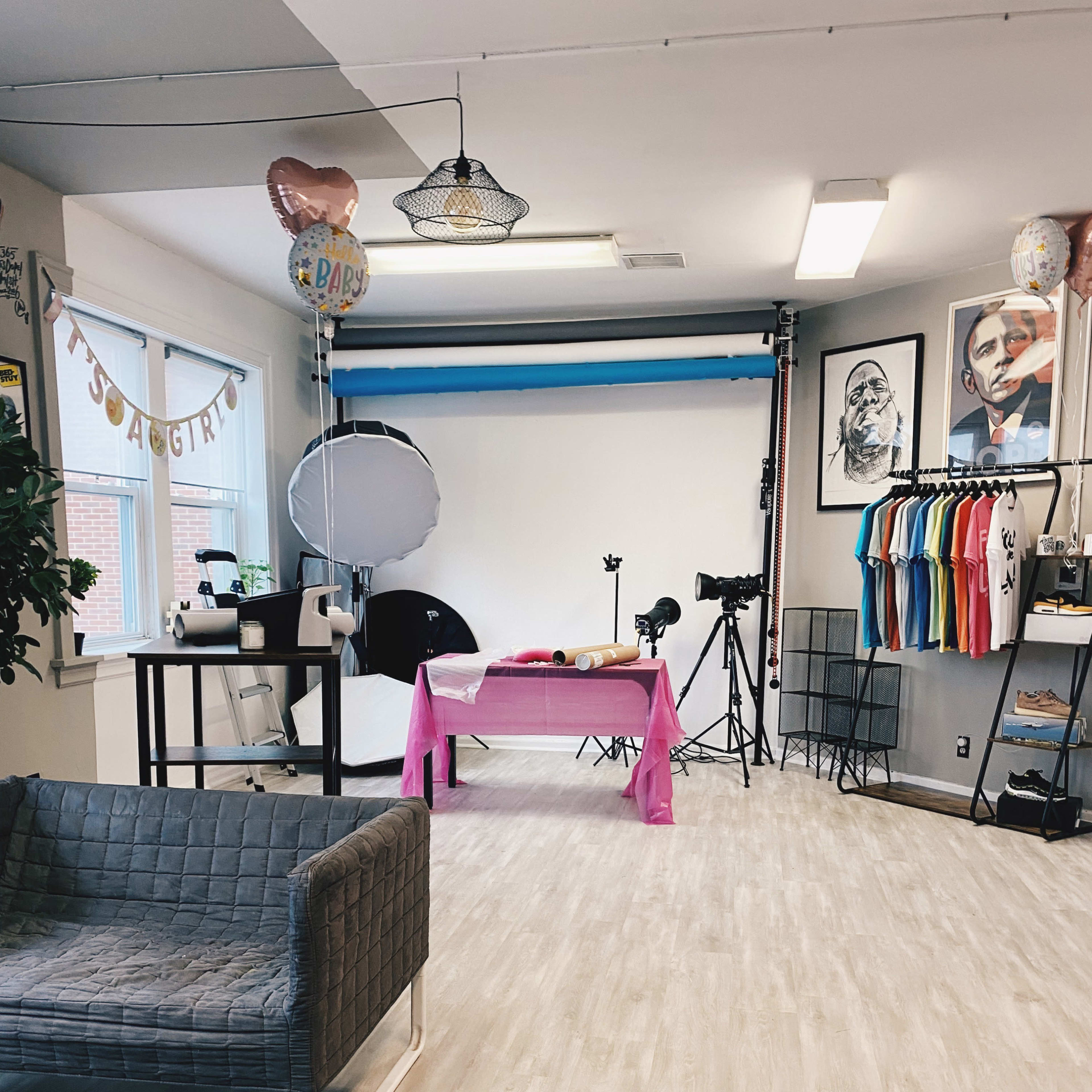 A studio space with a pink tablecloth, photography equipment, a gray couch, and colorful shirts hanging on one wall.