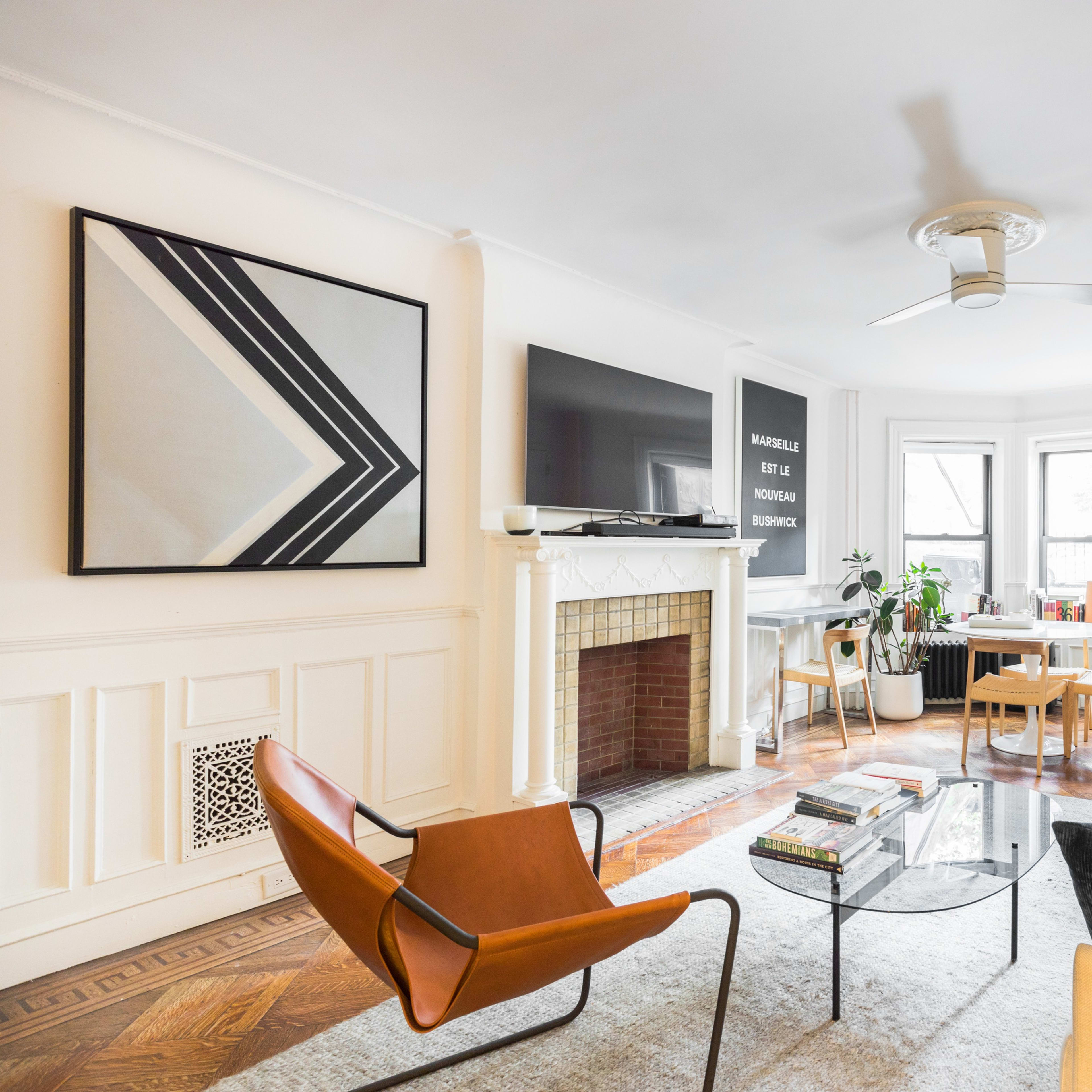 A modern living room features a beige sofa, an orange chair, a glass coffee table, and large windows with natural light illuminating the space.