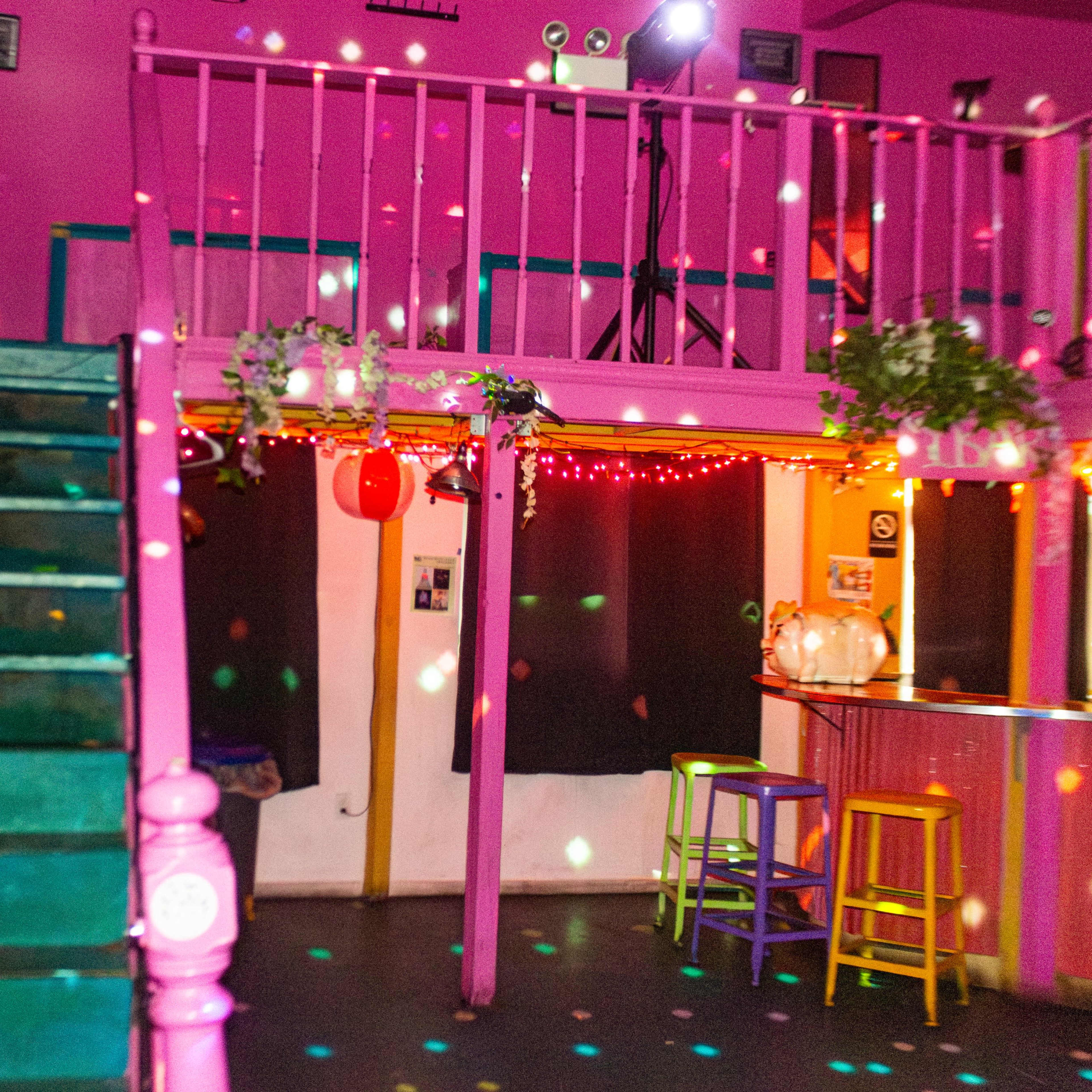 A vibrant indoor space with pink walls, a staircase leading to a second level, and colorful lights creating patterns on the floor.
