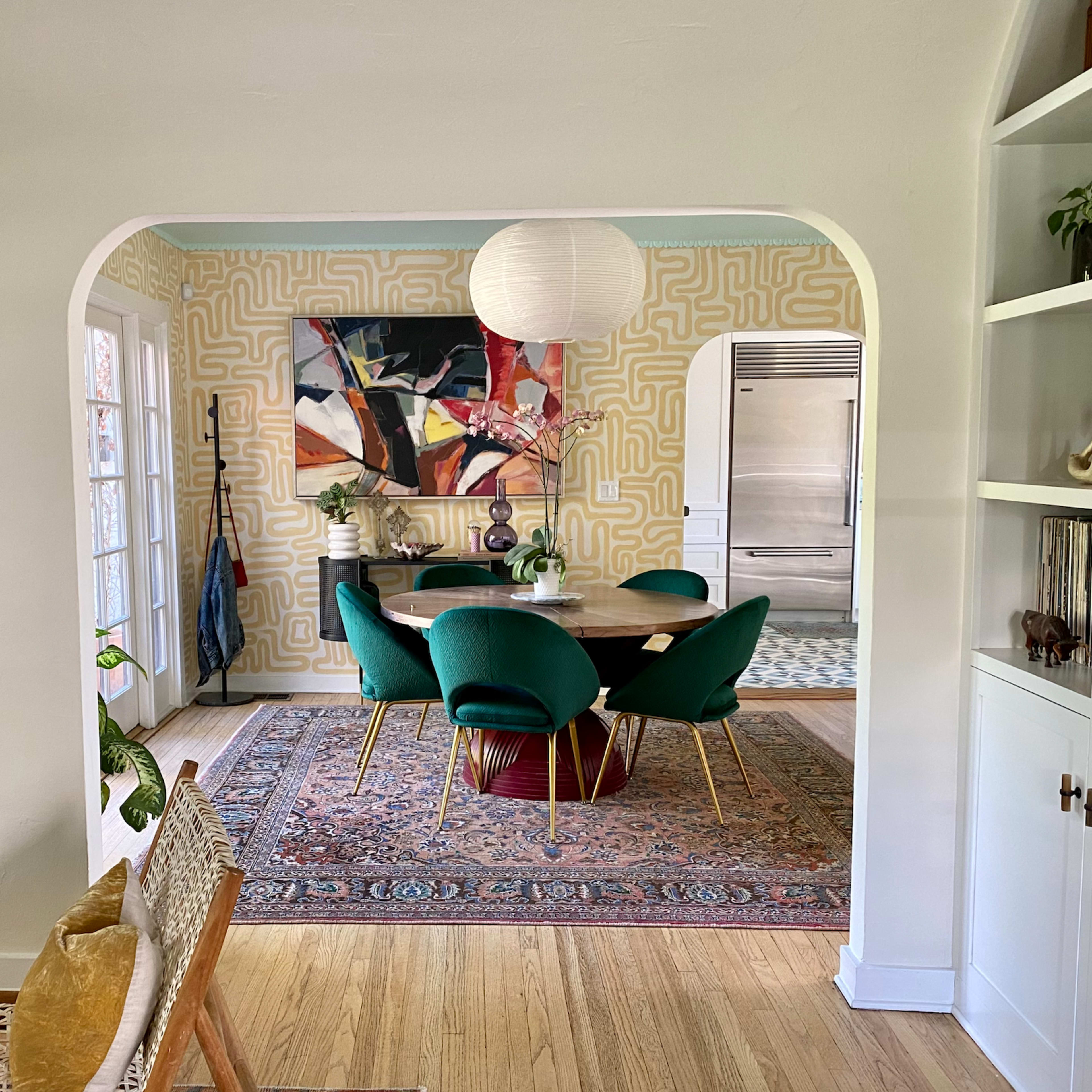 A dining area features a round table surrounded by green chairs, with abstract art on the walls, set against a patterned wallpaper and a decorative rug.