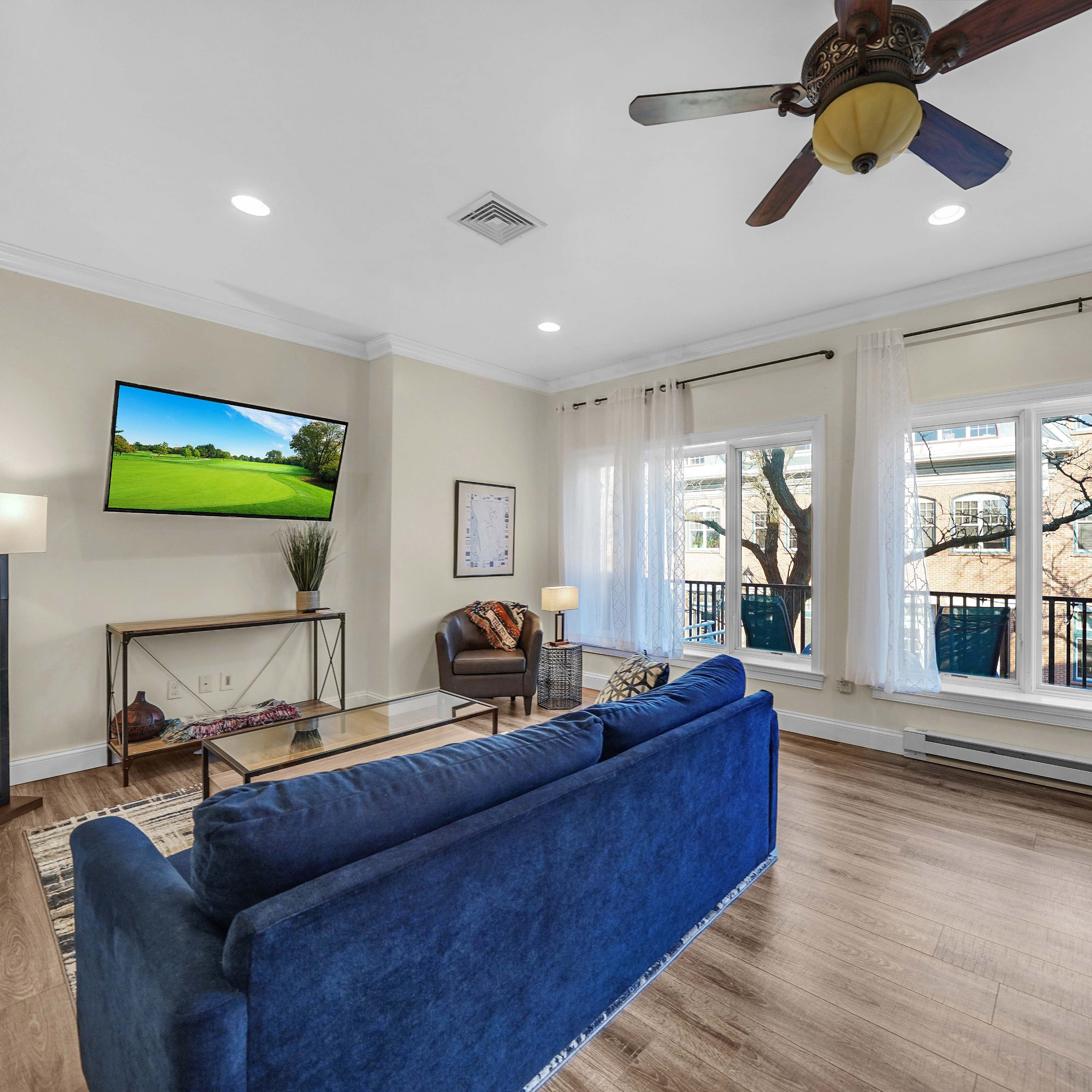 The living room features a blue sofa, a coffee table, and a television mounted on the wall, with large windows providing natural light.