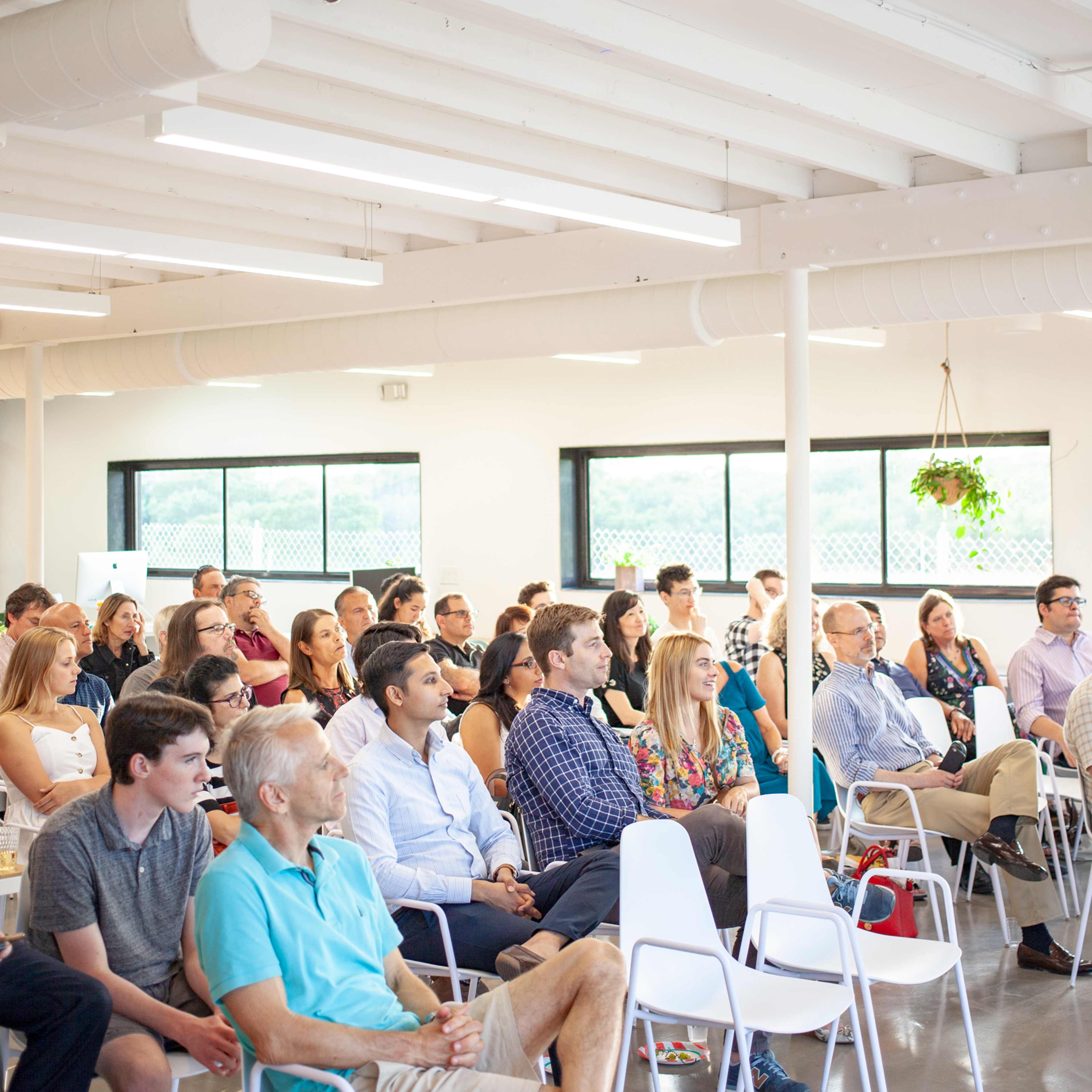 A diverse audience sits in a well-lit indoor space, attentively facing a speaker at the front.