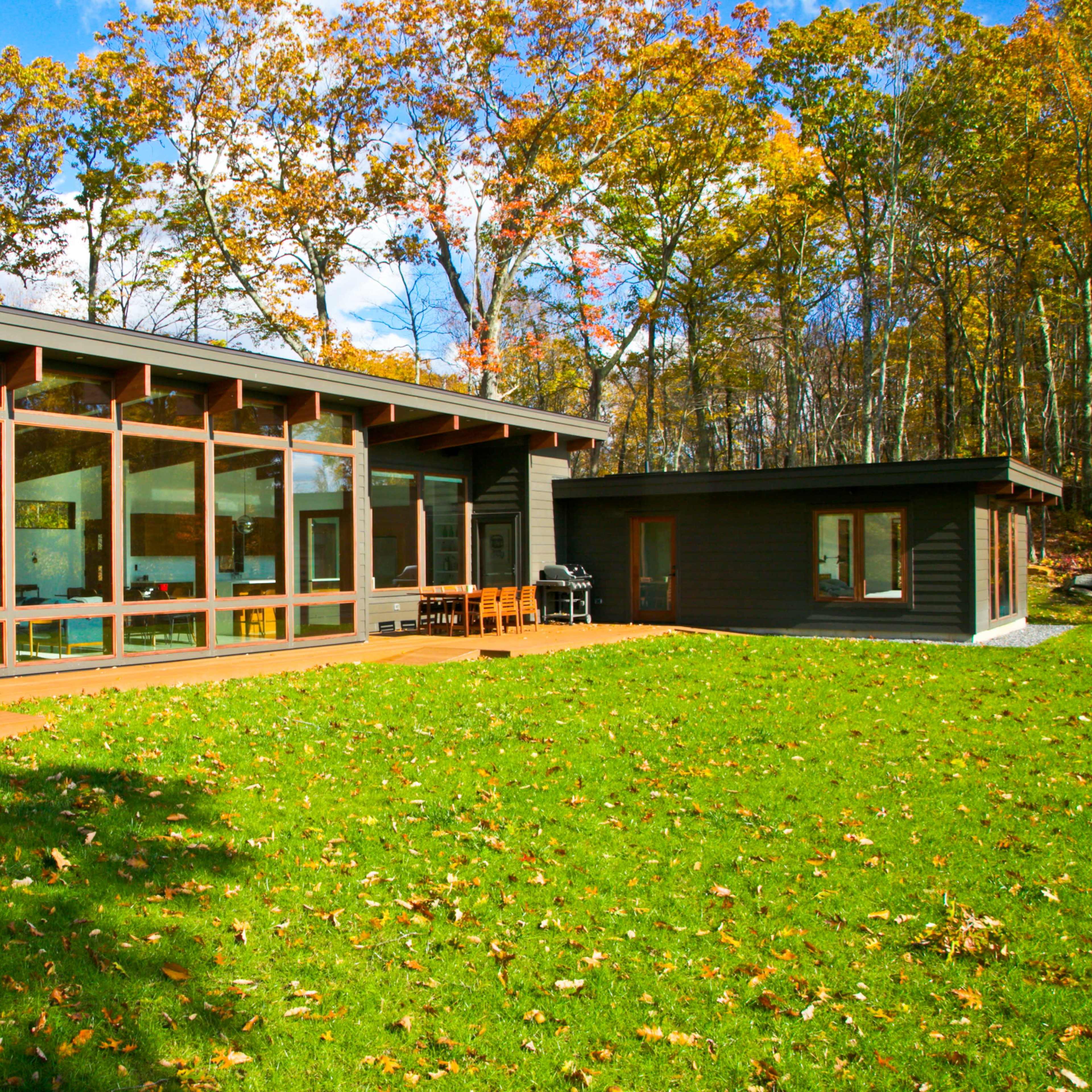 A modern house with large windows and a patio sits on a grassy yard surrounded by autumn trees.