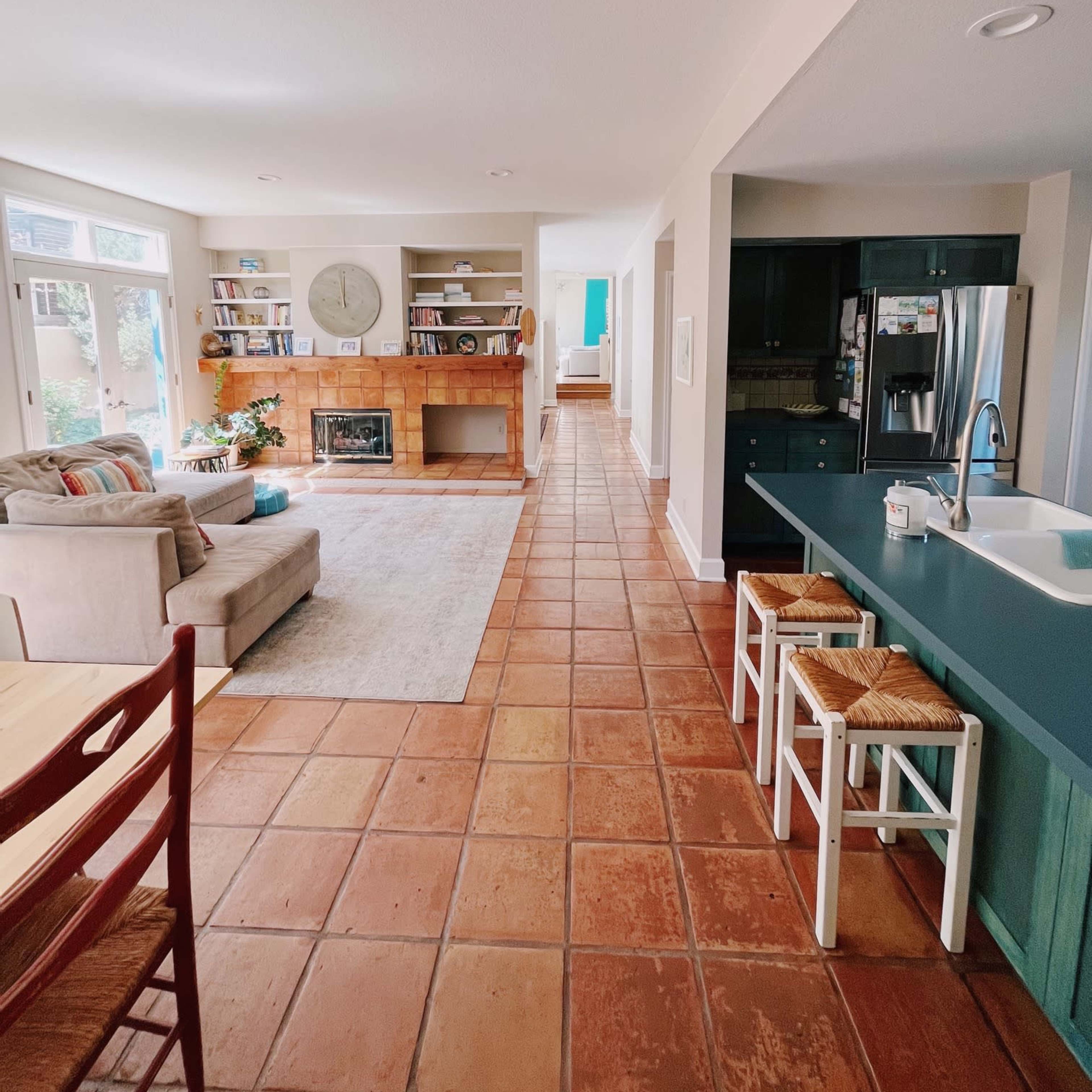 A spacious living area connected to a kitchen, featuring a tiled floor, a fireplace with built-in shelves, and a hallway leading to another room.