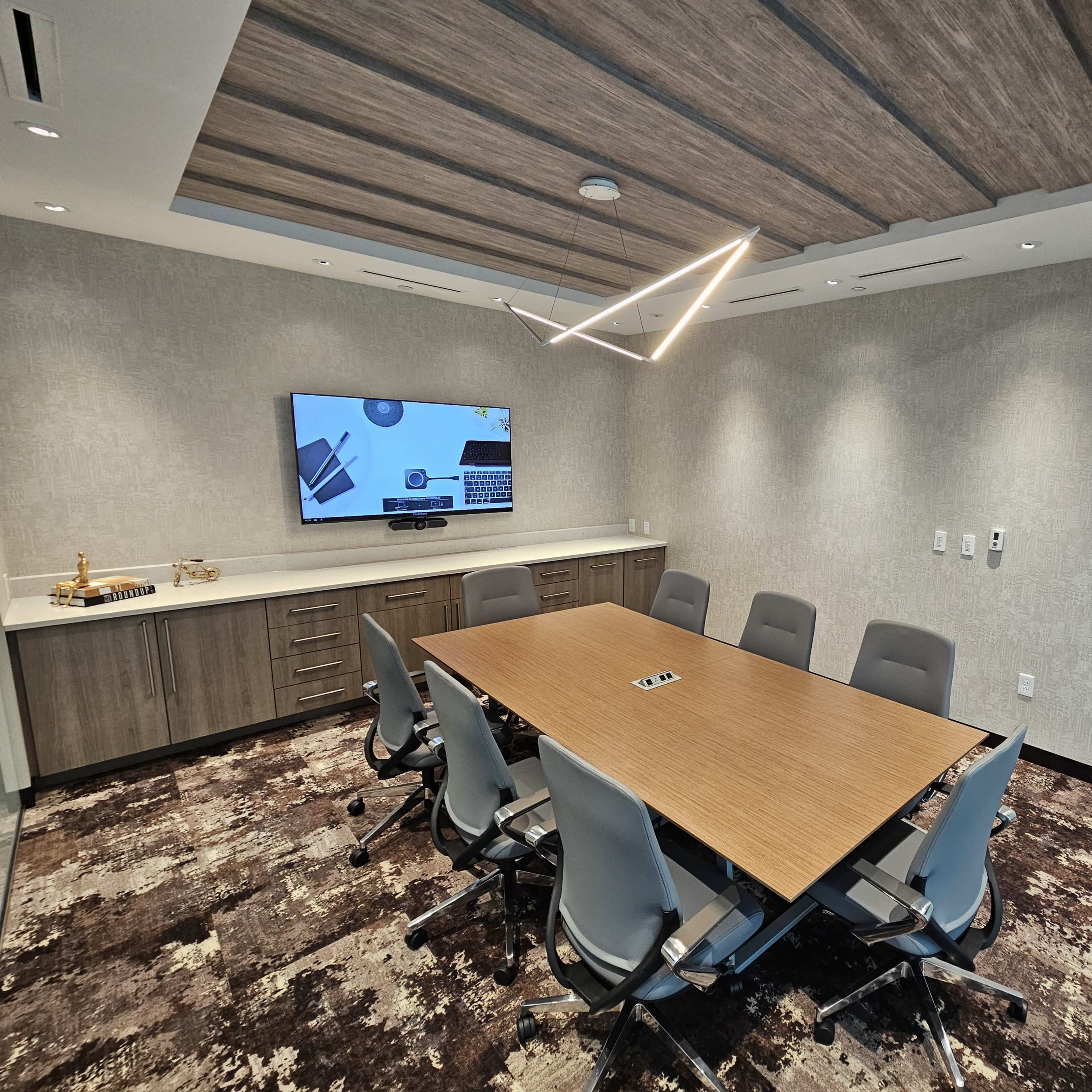 A modern conference room featuring a large wooden table surrounded by six chairs, with a television on the wall displaying a presentation.