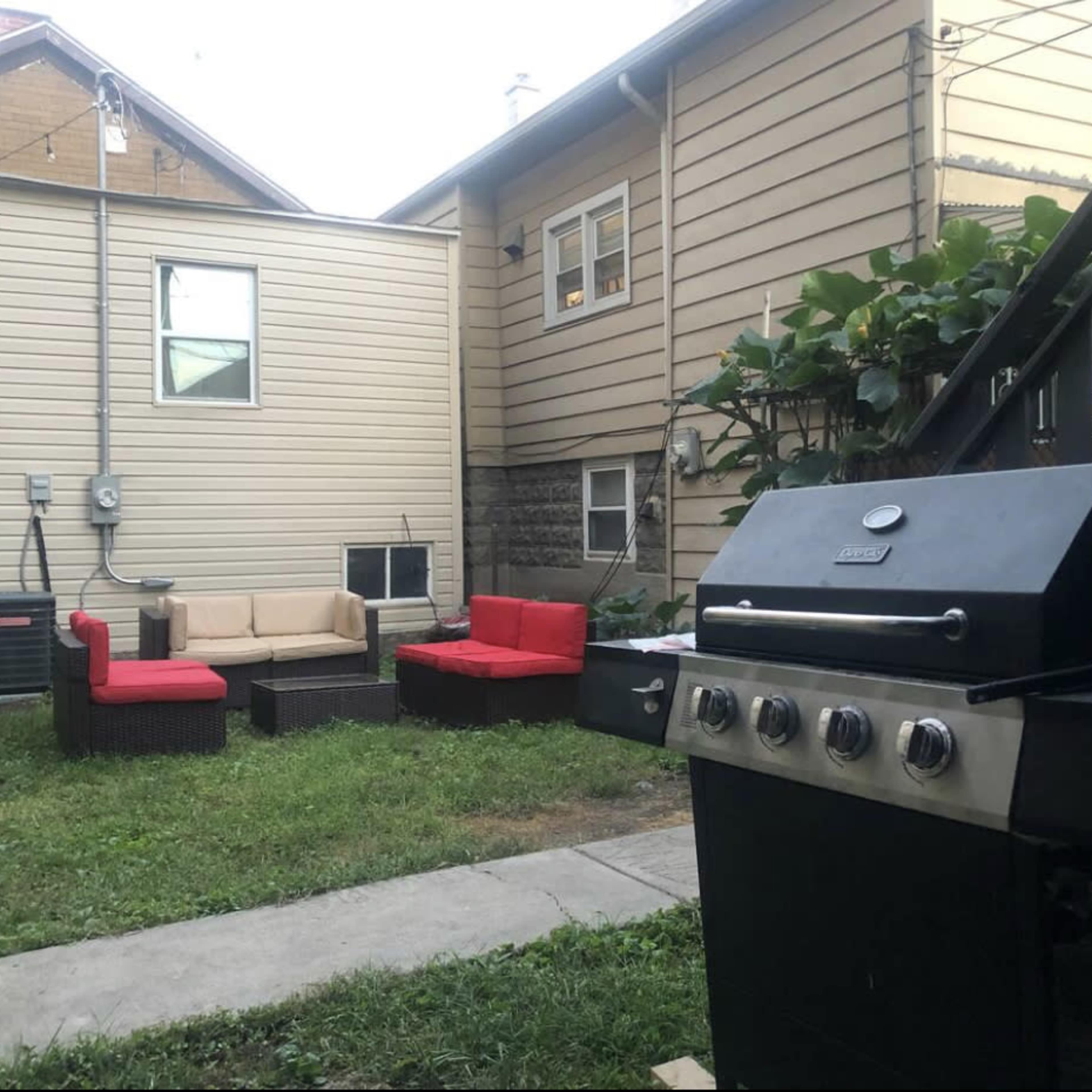 A backyard with a black grill in the foreground, red and beige outdoor seating, and a pathway leading to a house.
