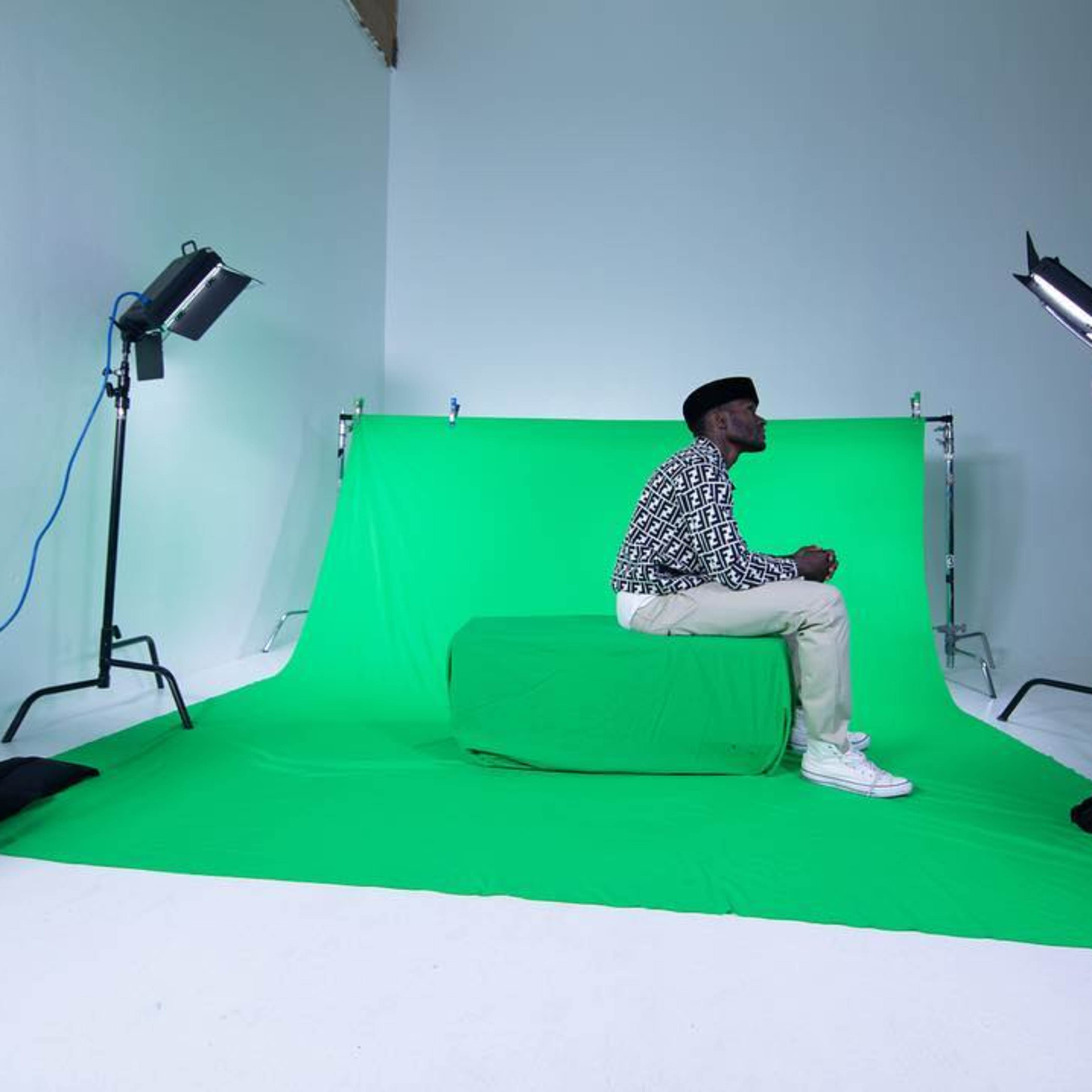 A man sits on a green set piece in a studio with a green backdrop, flanked by lighting equipment and a ladder.