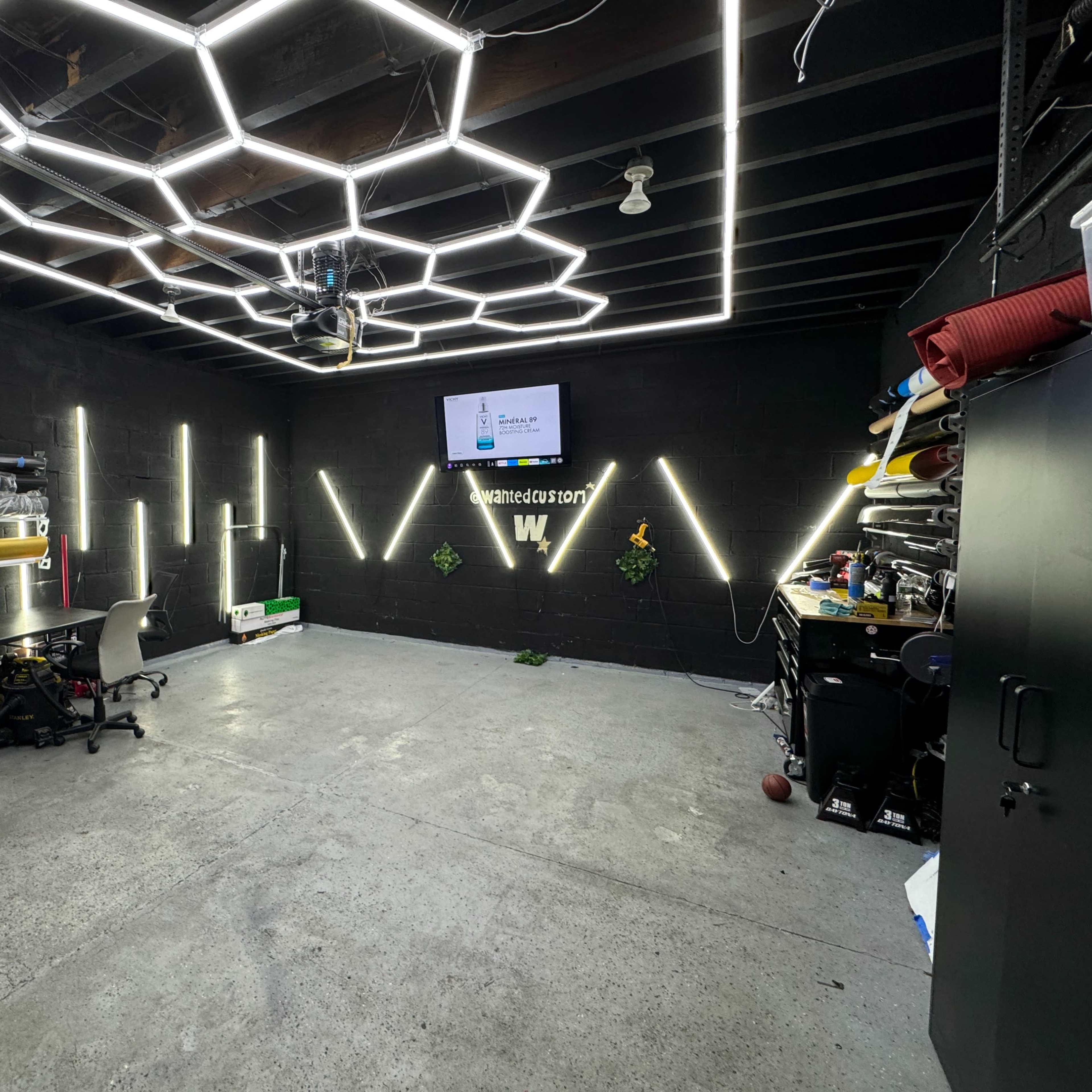 A modern garage or workshop space with hexagonal LED lighting, a television mounted on the wall, and various workstations and storage areas.
