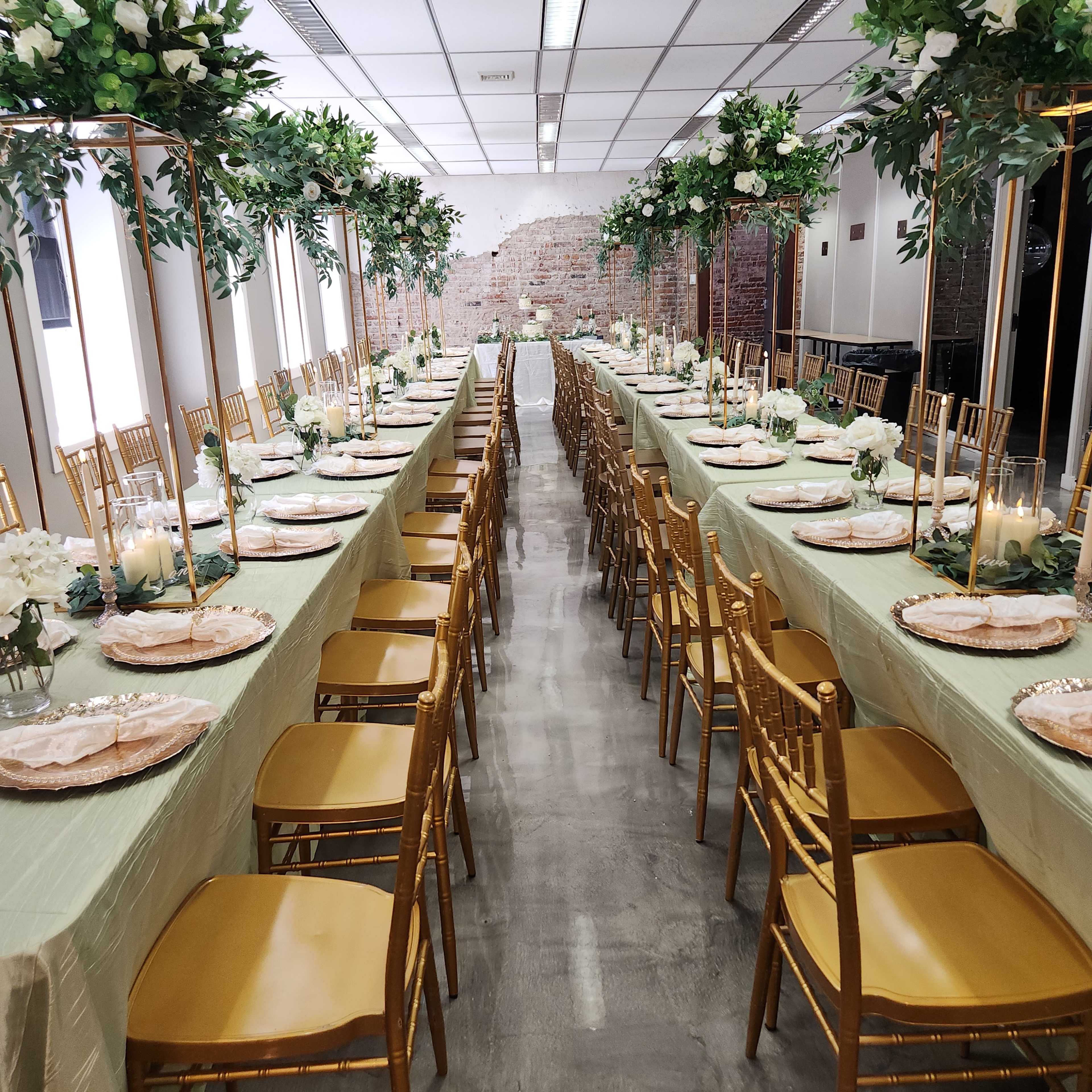 A long dining table is set with gold chairs, elegant tableware, and floral arrangements, surrounded by greenery and a brick wall backdrop.