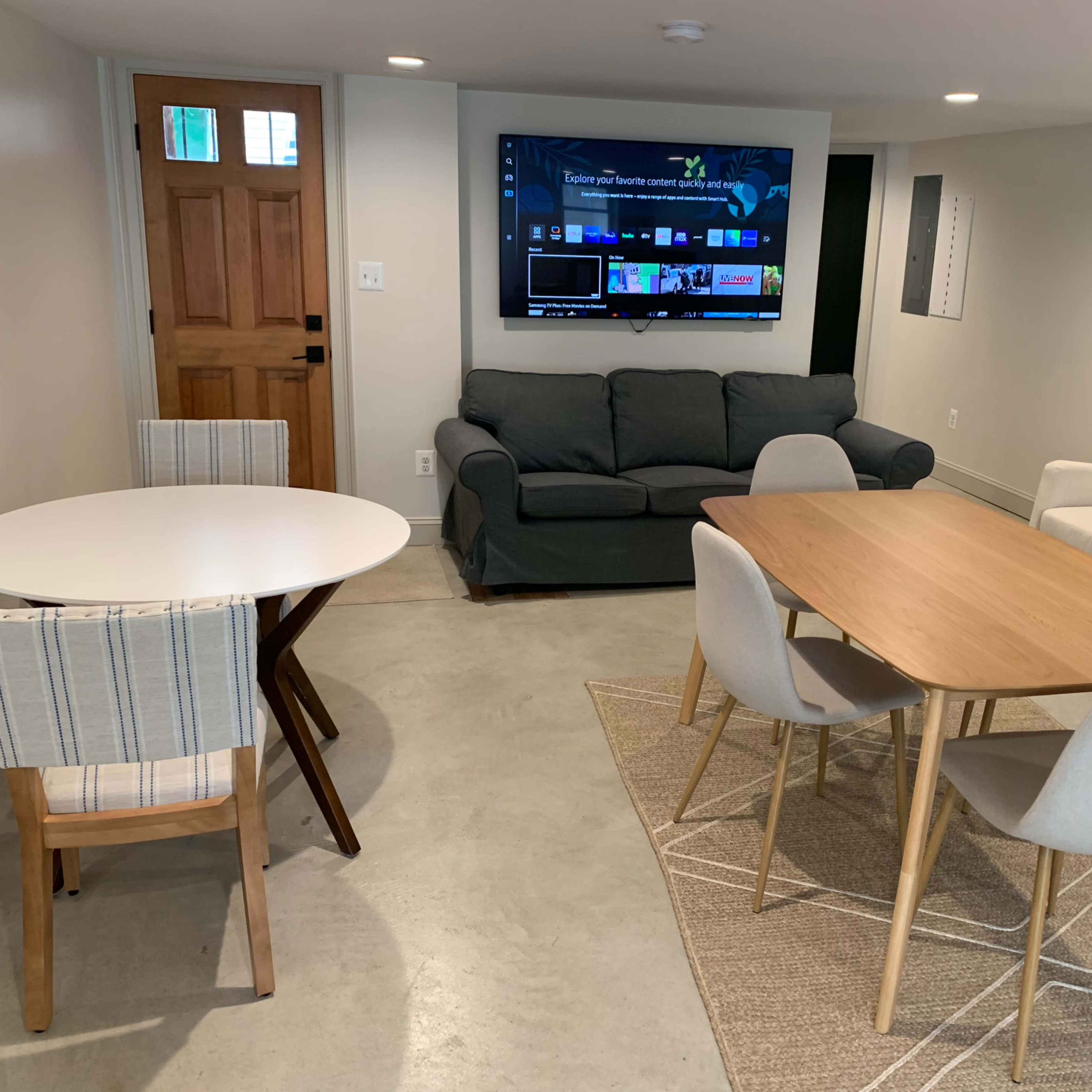 A modern basement living area featuring two seating areas, a television, a round table with striped chairs, and a rectangular table with light-colored chairs.