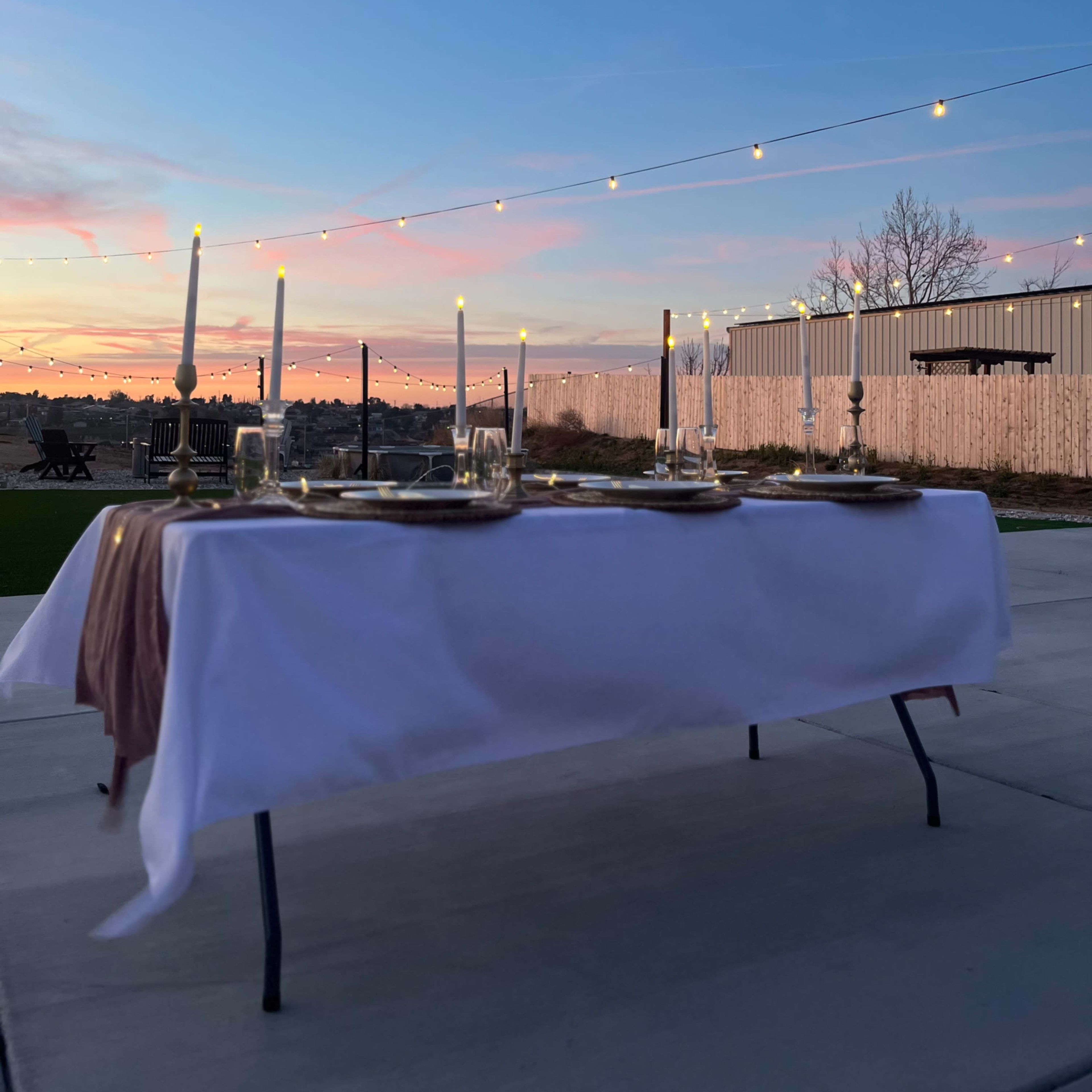 A table set with candles and dinnerware is positioned outdoors under a colorful sunset and string lights.