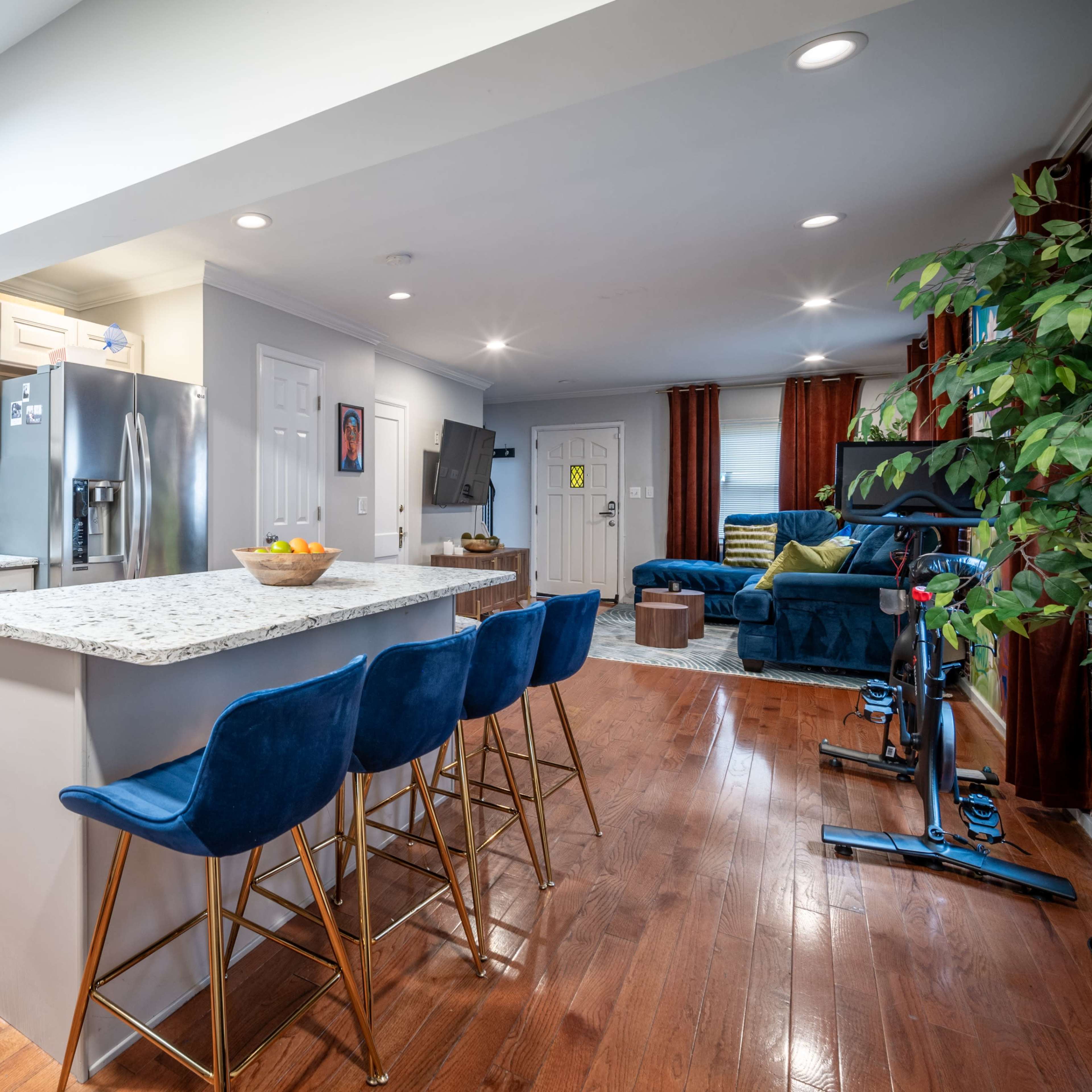 A modern kitchen features a large island with blue bar stools, granite countertops, and stainless steel appliances, illuminated by overhead lights.