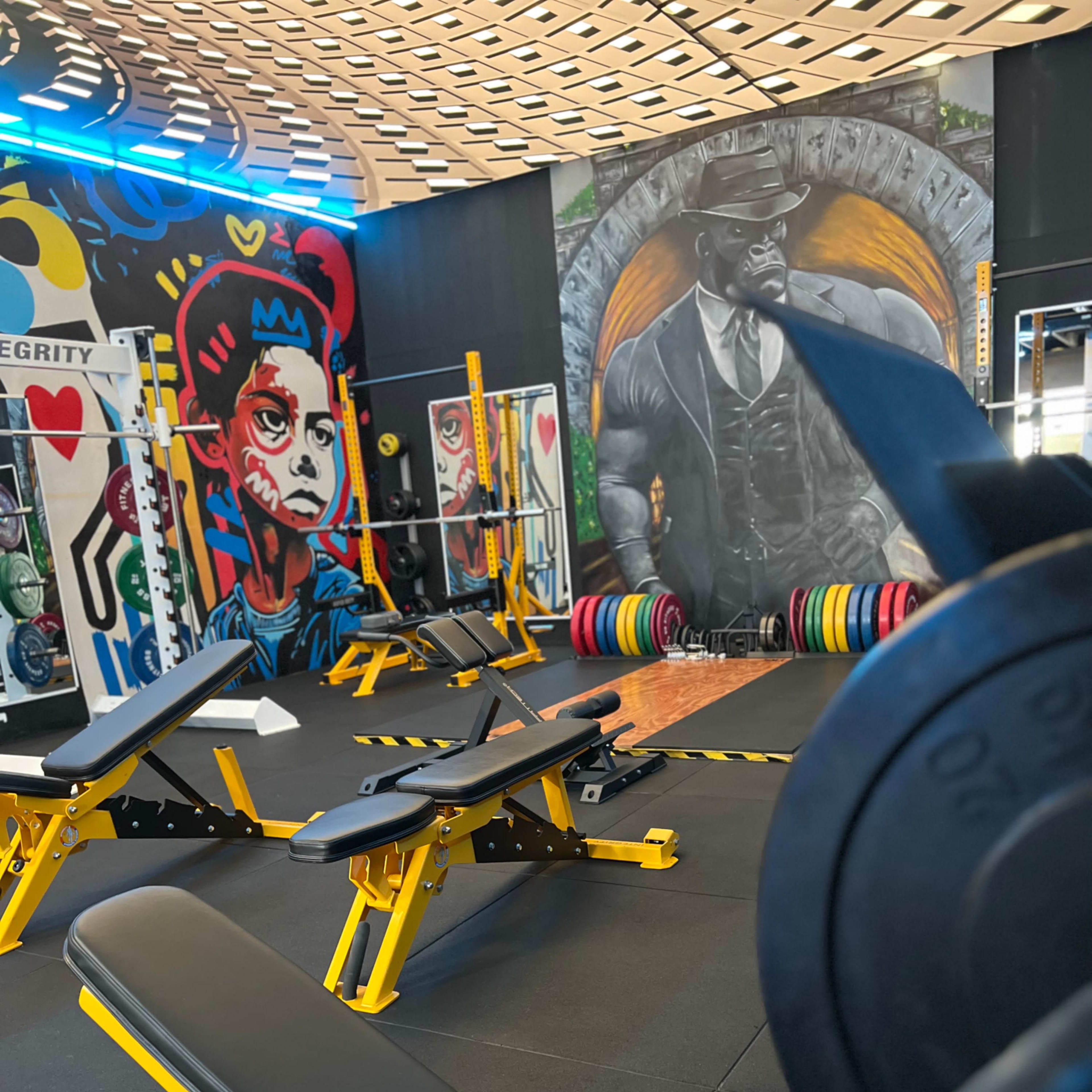 The gym features weight benches and a colorful mural of a muscular figure alongside various weight plates.