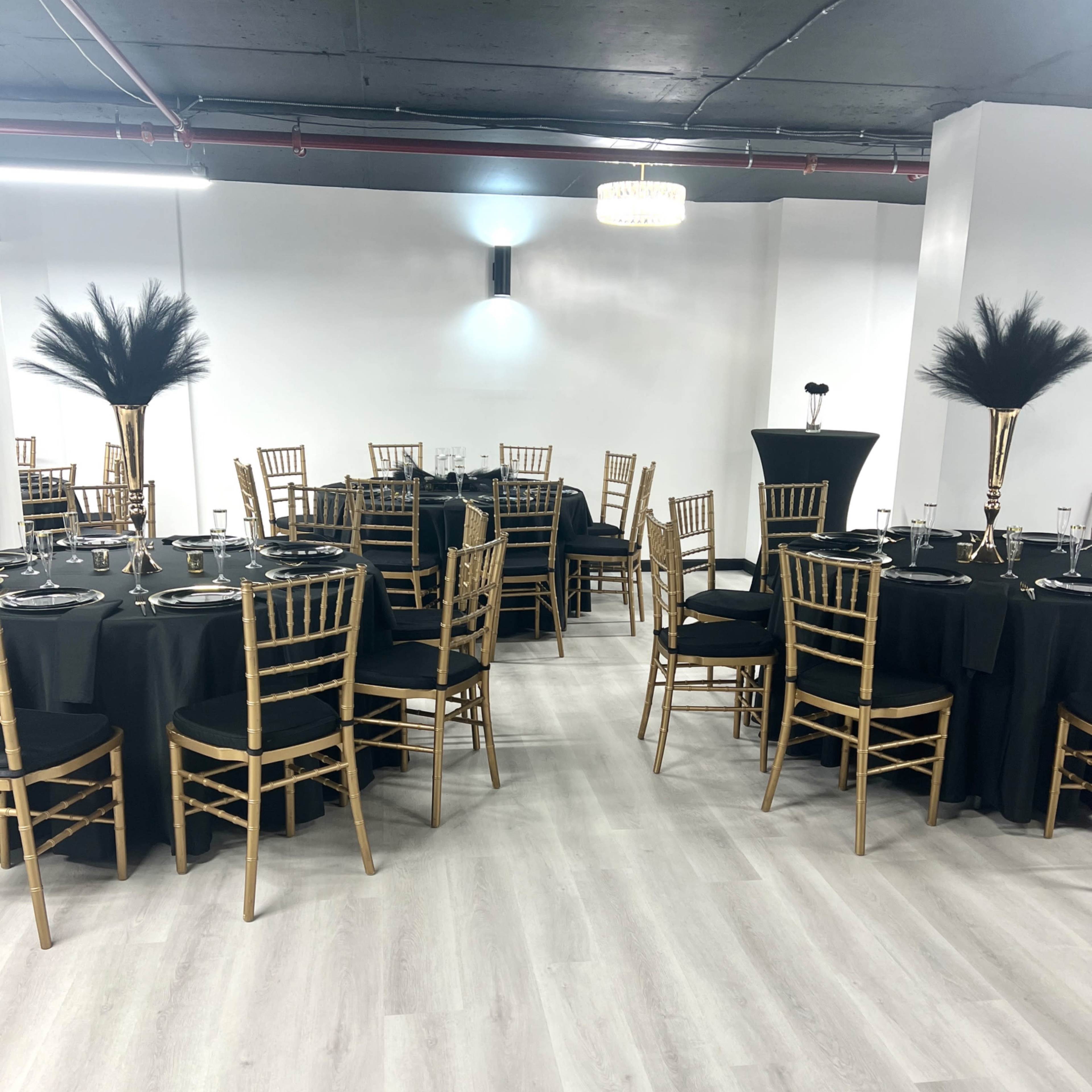 A well-arranged event space with round tables covered in black cloth, gold chiavari chairs, and centerpieces featuring tall, feather arrangements.