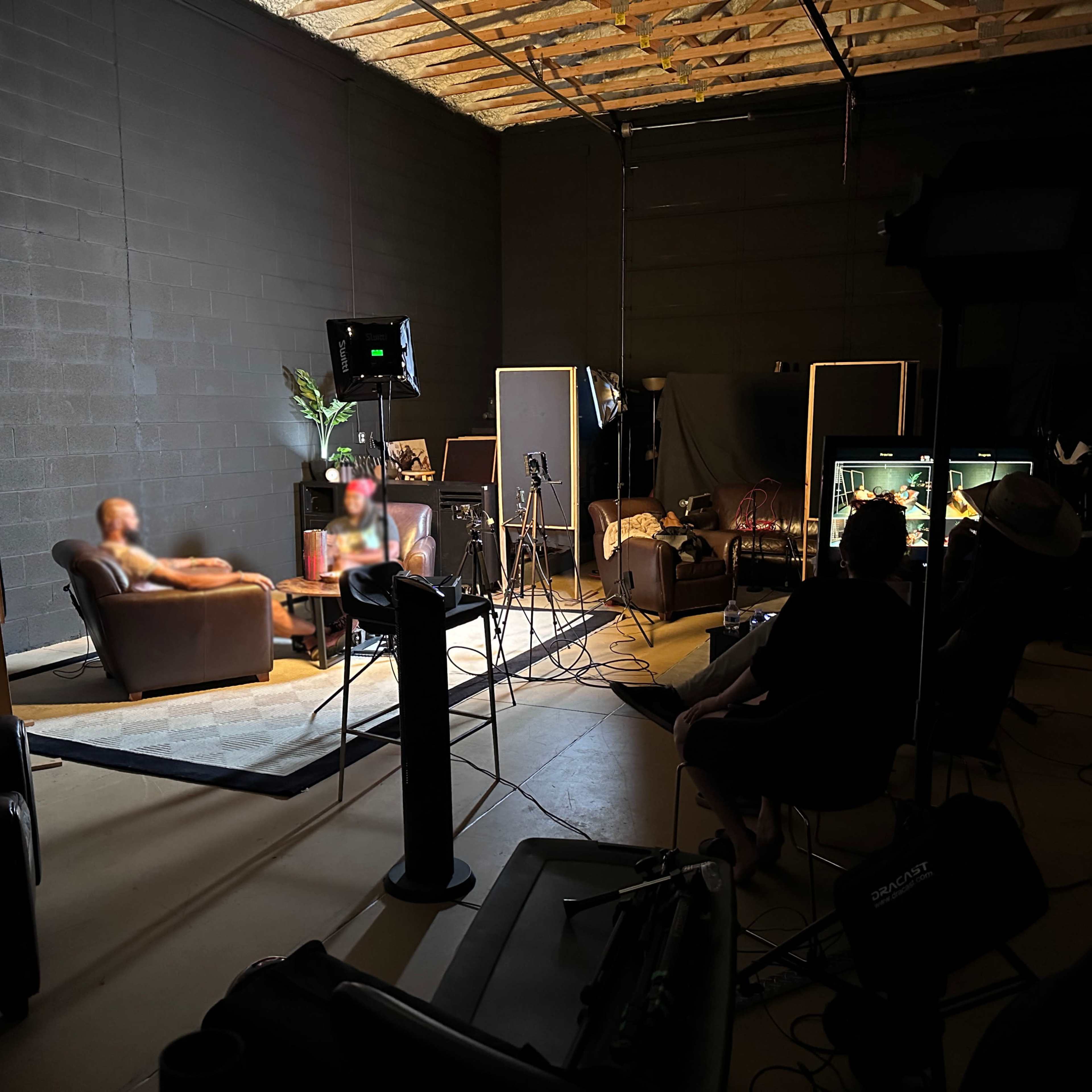A dimly lit studio with two people sitting on a couch, surrounded by cameras and lighting equipment.
