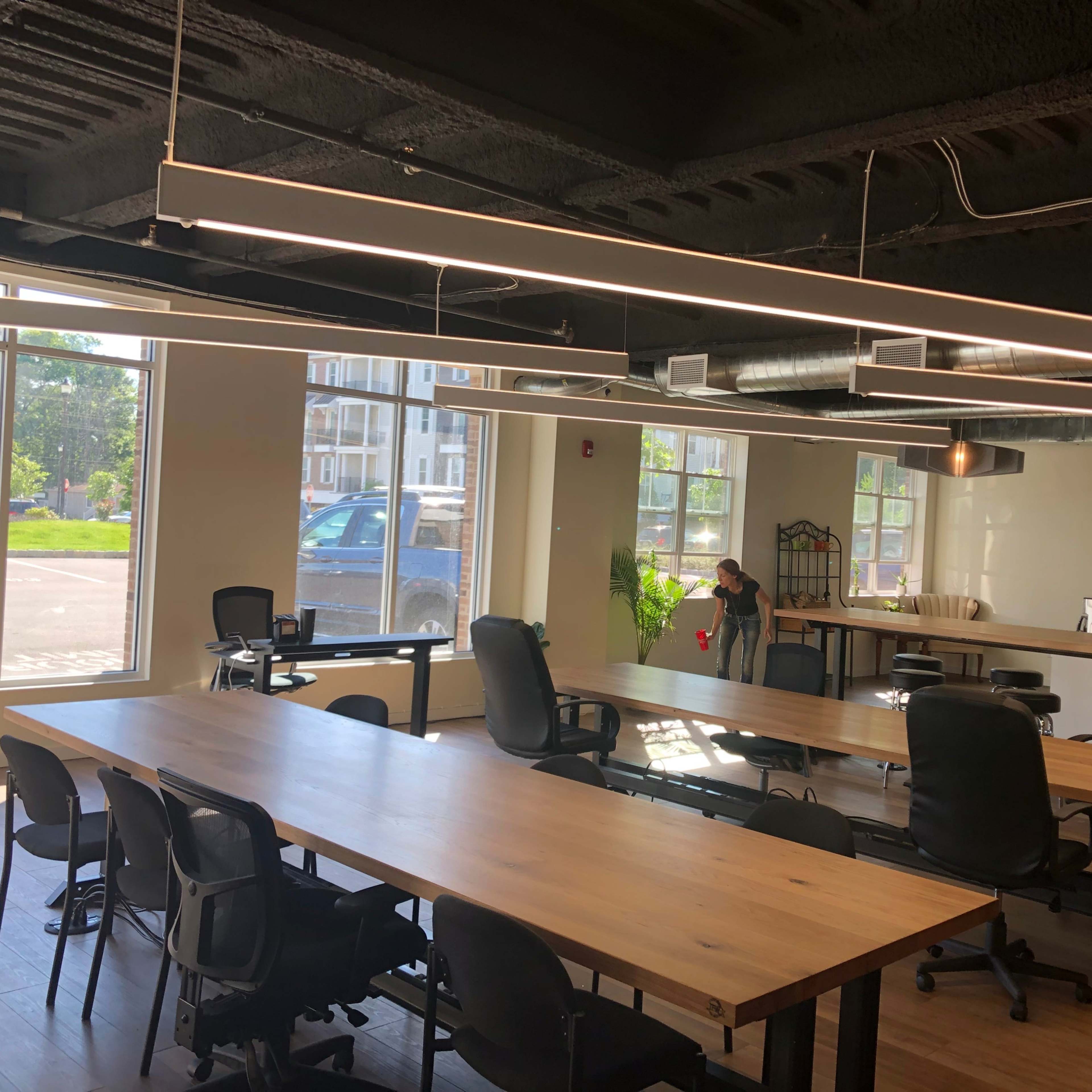 An empty modern office space features large windows, wooden tables, and ergonomic black chairs, with a person cleaning in the background.