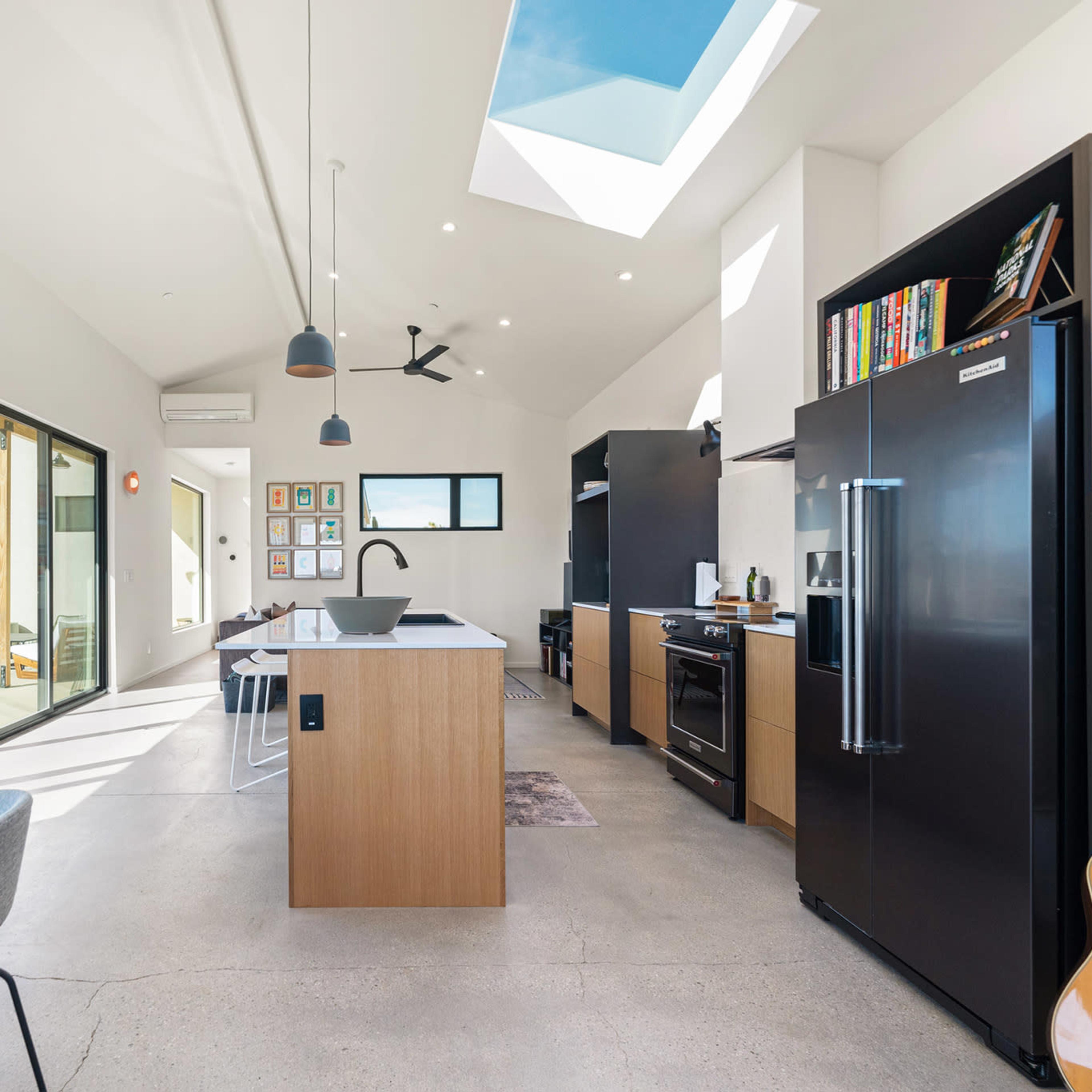 A modern, open-concept kitchen and dining area with large windows, a skylight, and sleek black appliances.