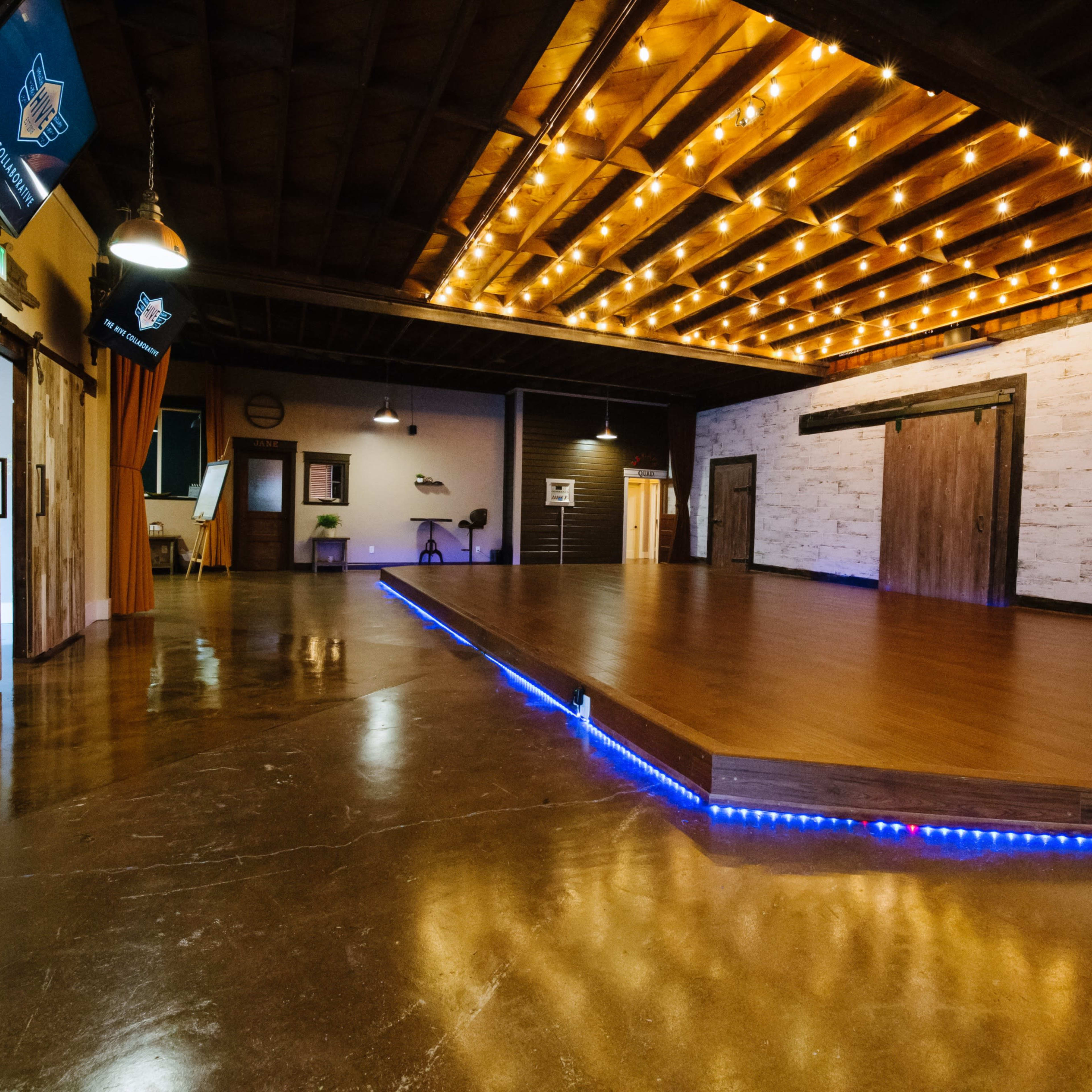 A spacious room with polished concrete floors, a wooden stage outlined by blue LED lights, and warm string lights along the ceiling.