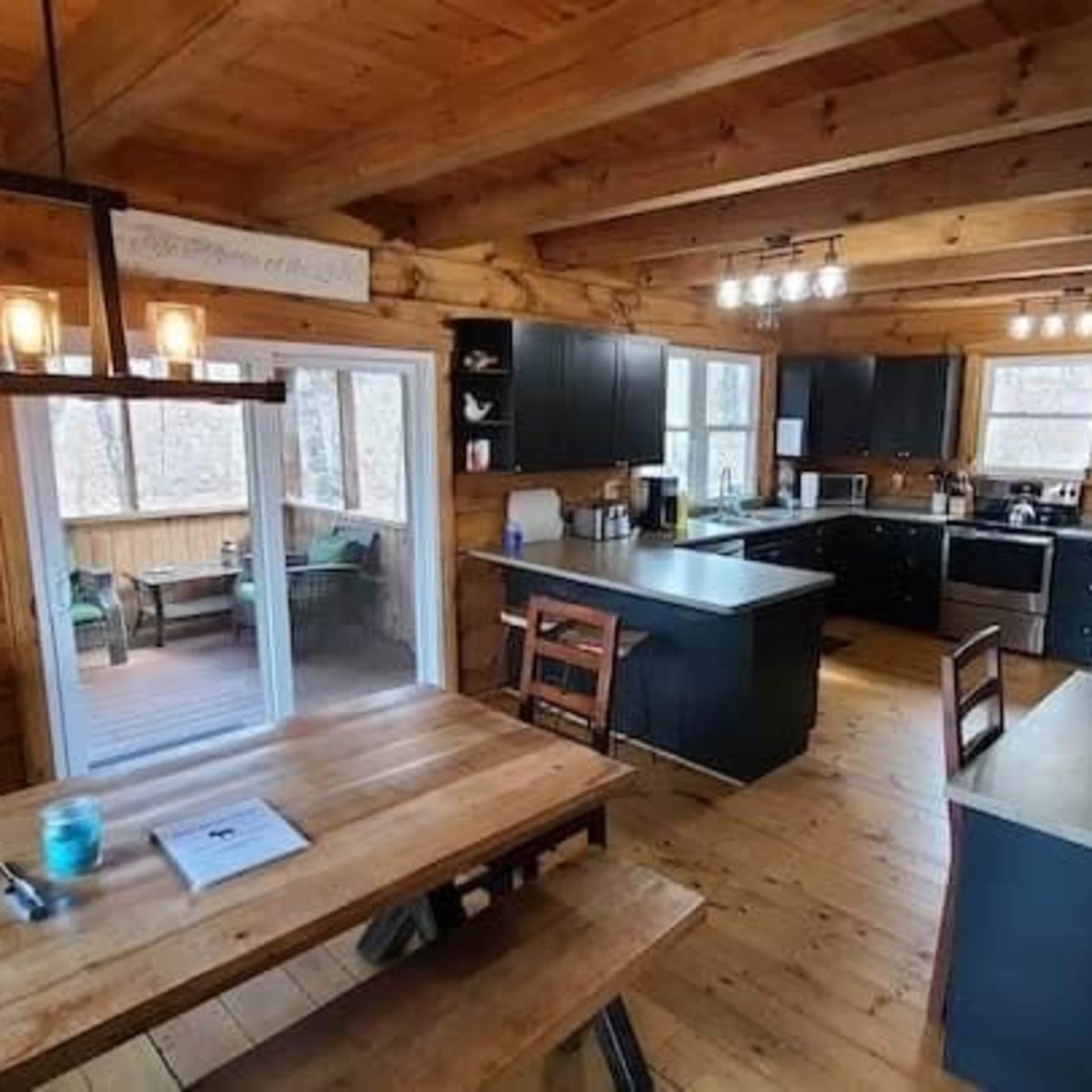 A rustic kitchen with wooden beams, dark cabinets, a central island, and a dining table, featuring large windows that overlook a deck.