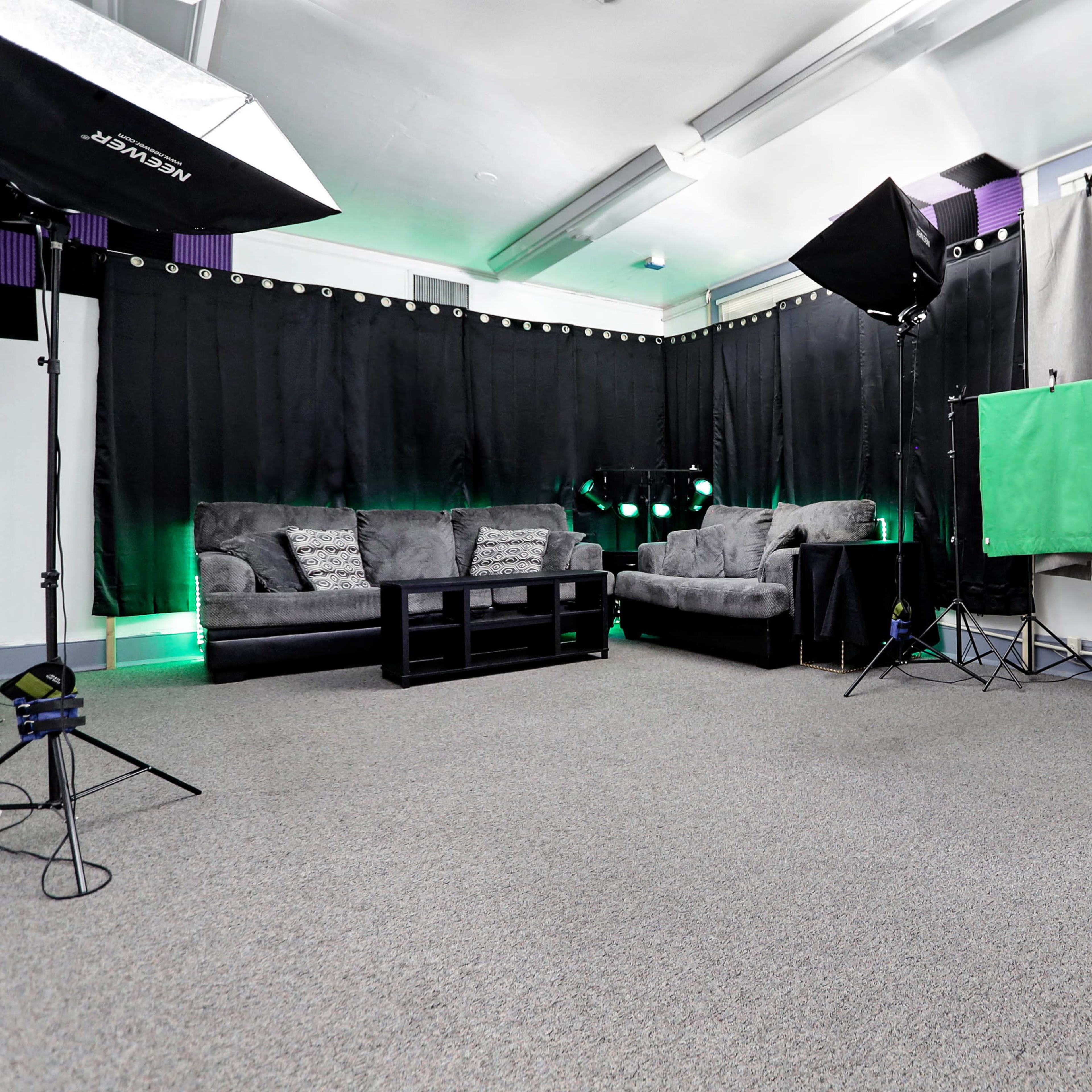 A studio setup with two gray and one green backdrop on stands, a sofa set, and lighting equipment in a carpeted room.