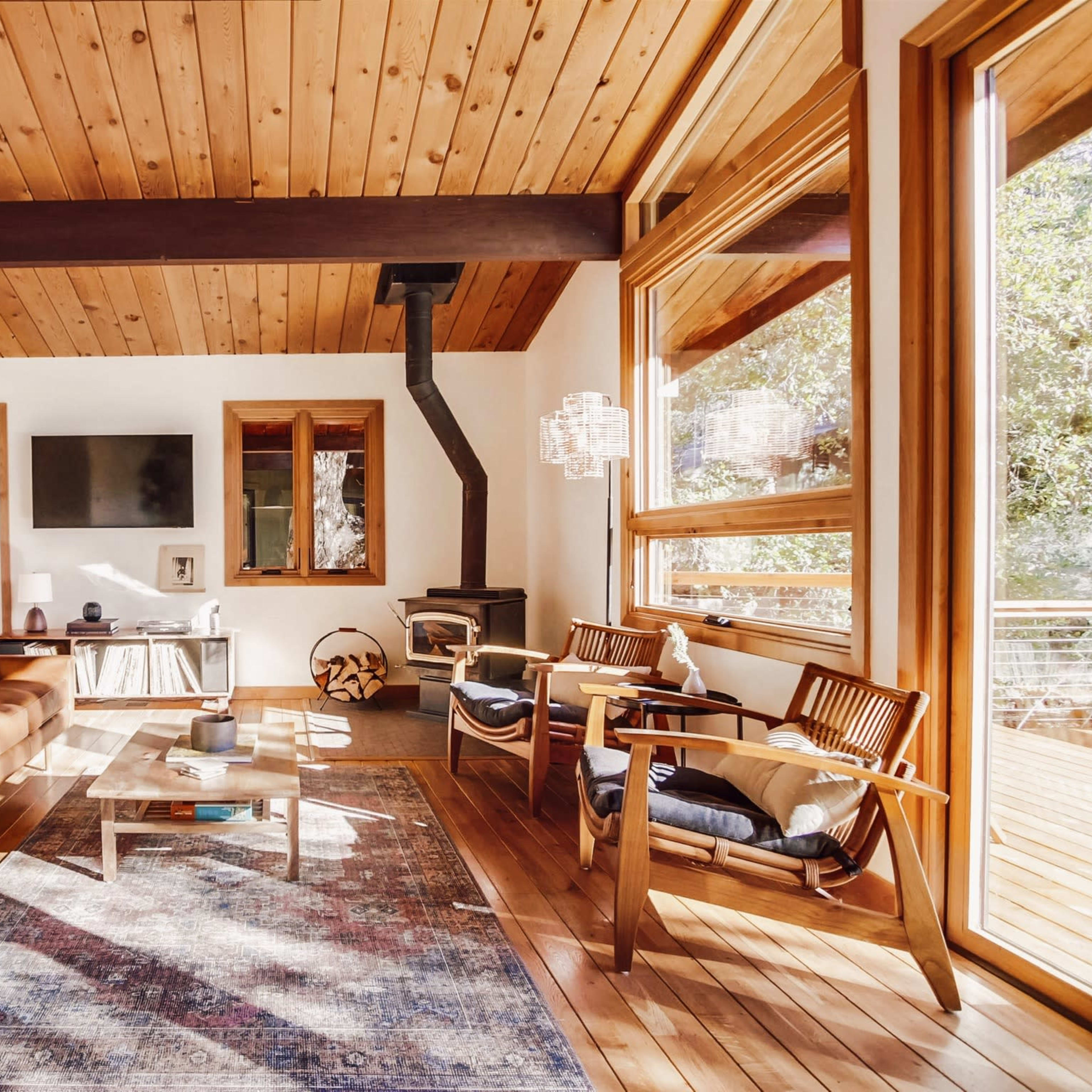 A spacious living room features a brown leather sofa, wooden flooring, a stone fireplace, and large windows allowing natural light to illuminate the space.