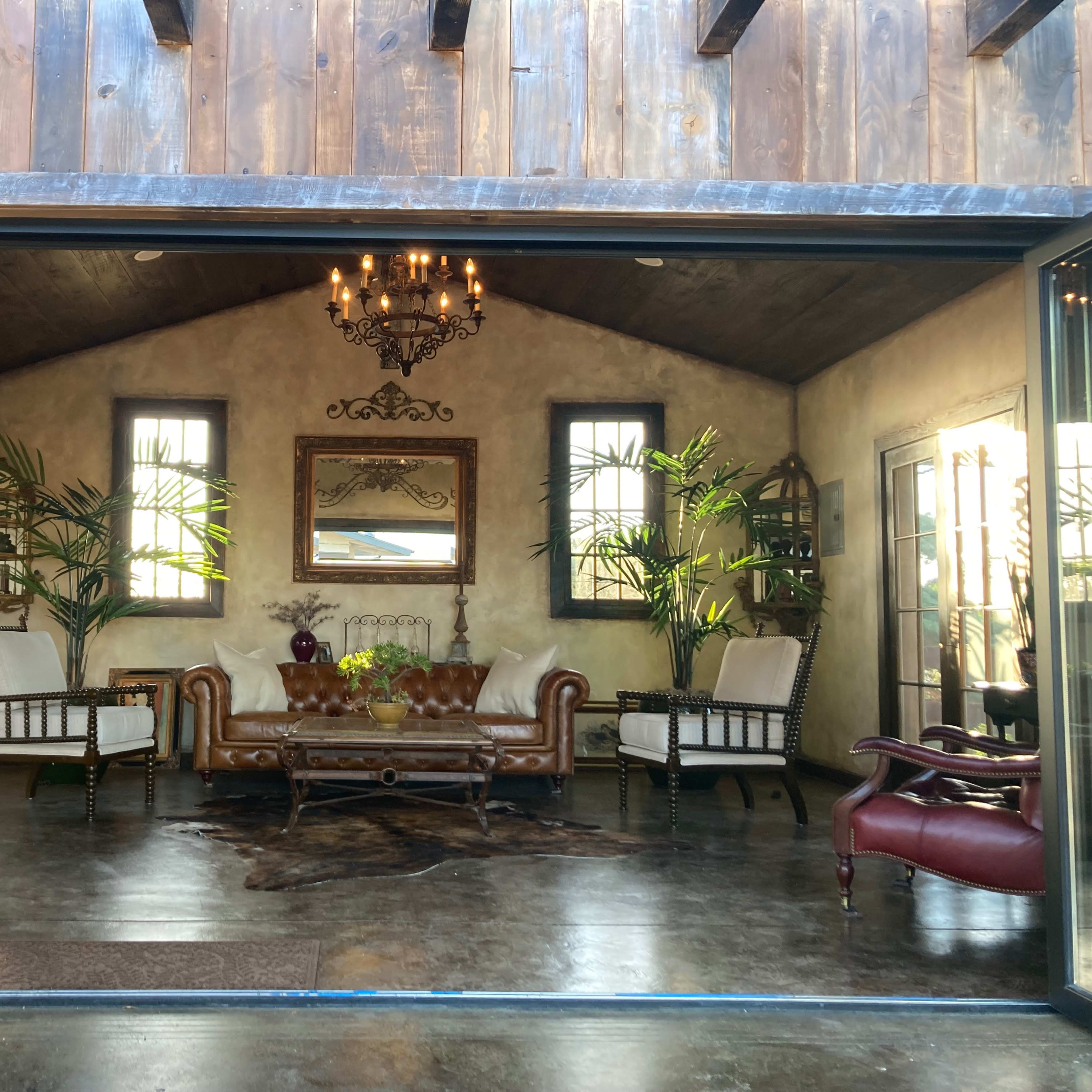 A cozy interior with a brown leather sofa, two white armchairs, a coffee table, and decorative plants under a chandelier, framed by large glass doors.