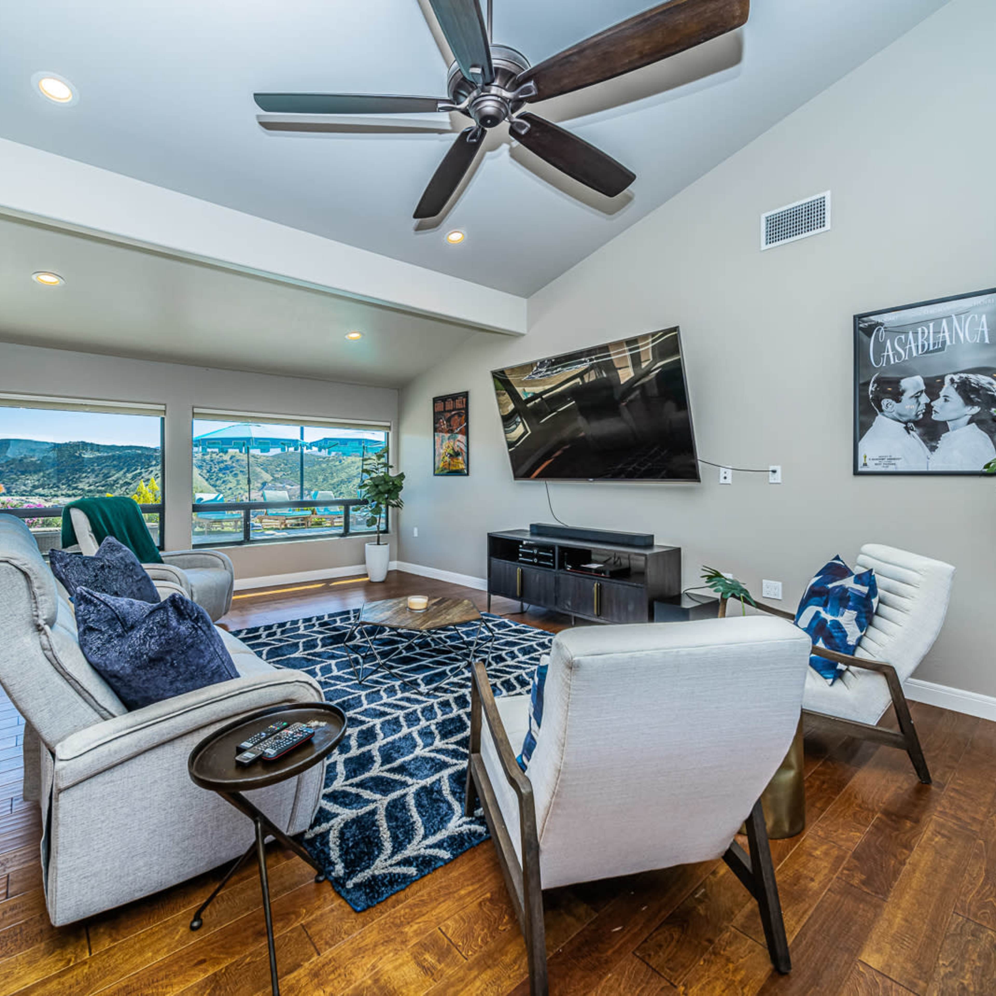 A modern living room features a large television, comfortable seating, and panoramic views of the mountains through large windows.
